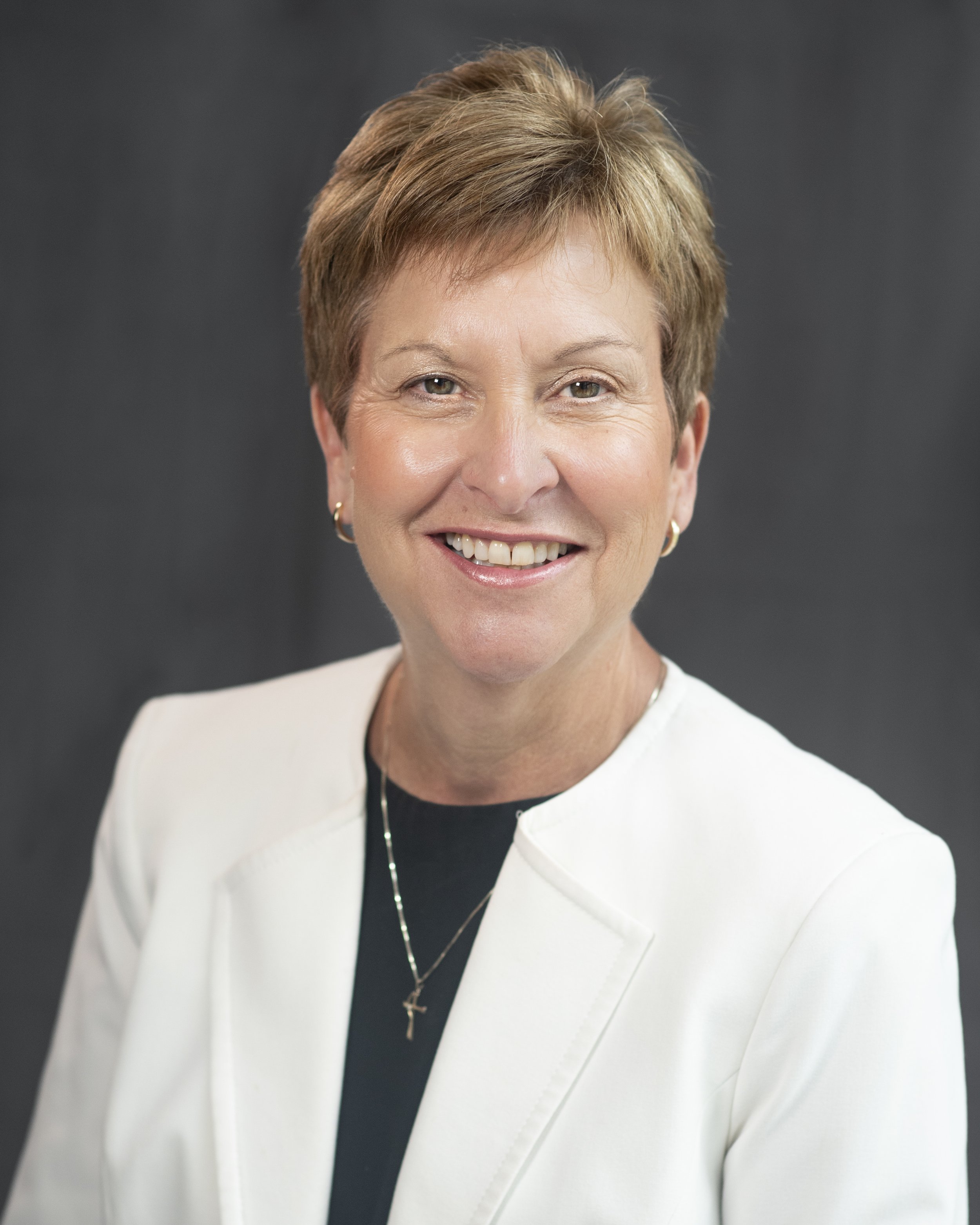 Portrait of a middle-aged woman smiling, wearing a black top and a gold necklace, inside a wooden-paneled room with warm lighting.