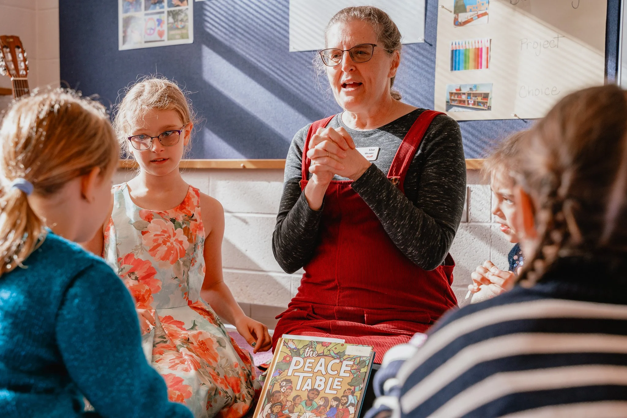 A teacher or adult woman with glasses and a red apron sitting on the floor with children in a classroom, holding her hands together as she speaks to them, with children sitting around her and a book titled 'The Peace Table' in the foreground.