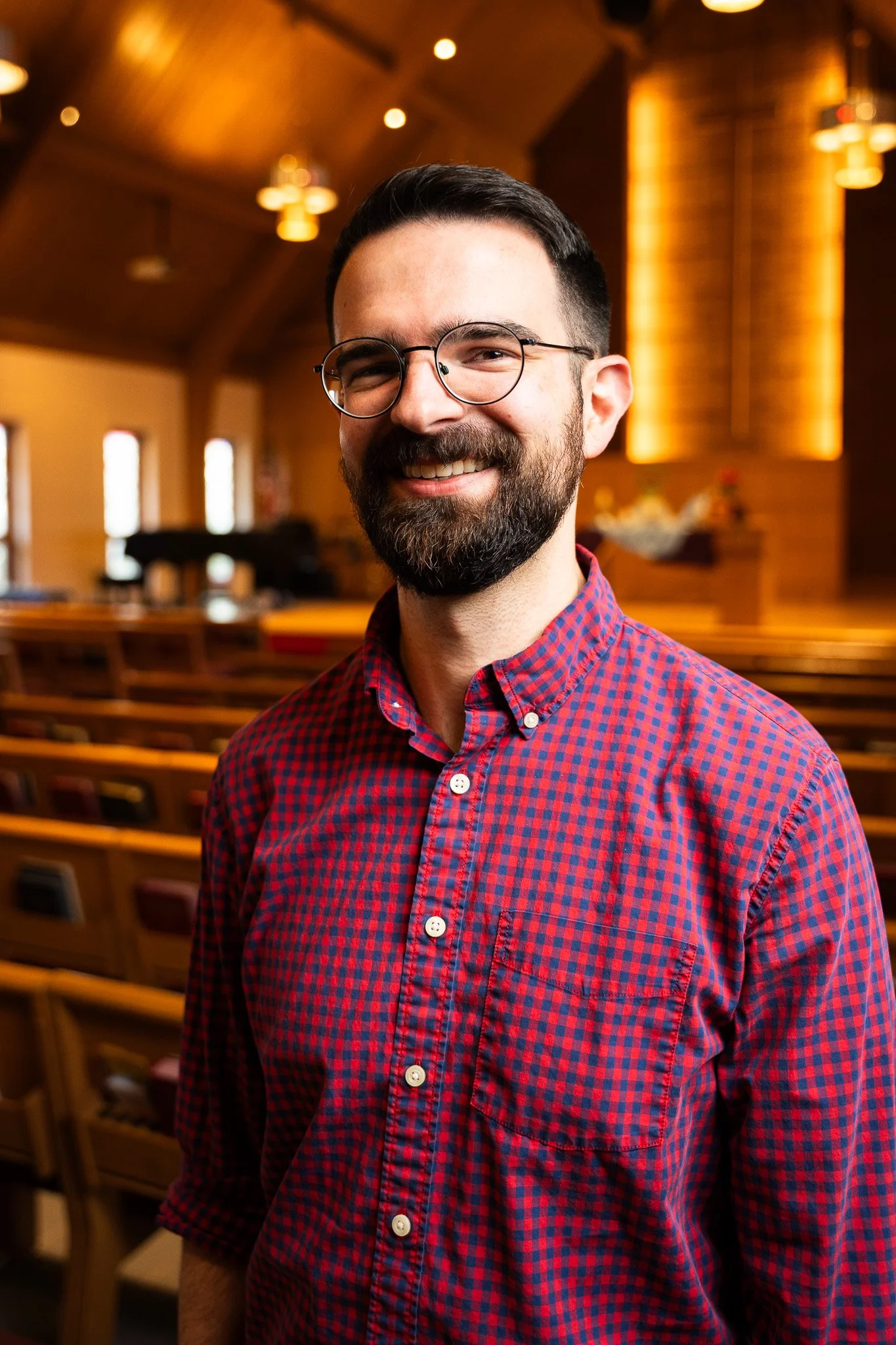 A smiling man with dark hair, glasses, and a beard wearing a red and blue checkered shirt standing inside a warmly lit church.