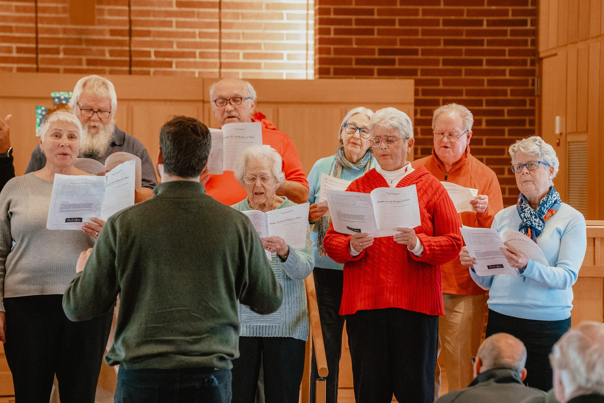 A choir of elderly people singing during a performance, led by a conductor, inside a wooden-paneled room.
