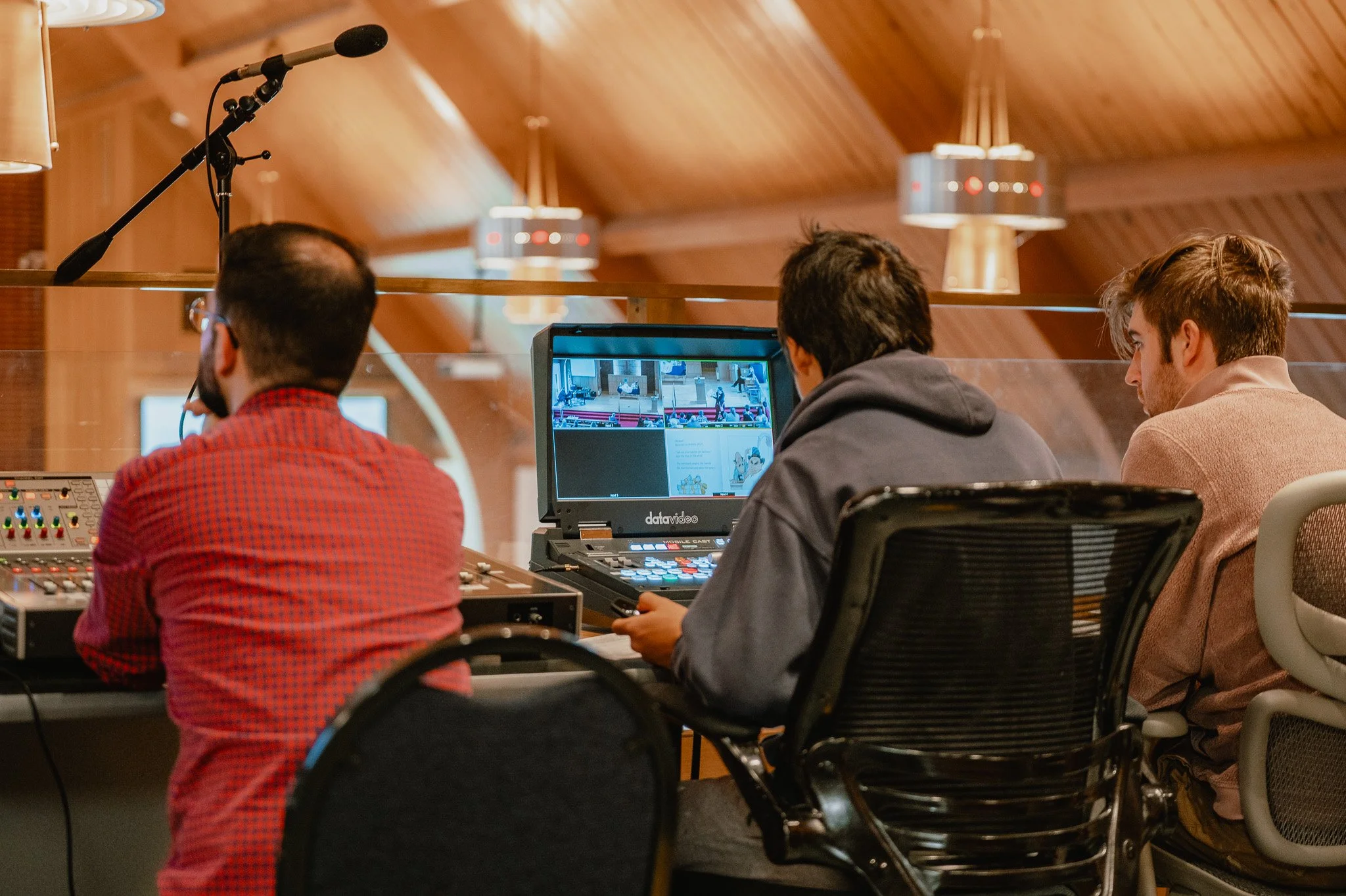 Three men sitting at a table in a recording studio or control room, with one using a laptop and sound mixing equipment, and a microphone hanging above them. They are watching video footage on the laptop screen.