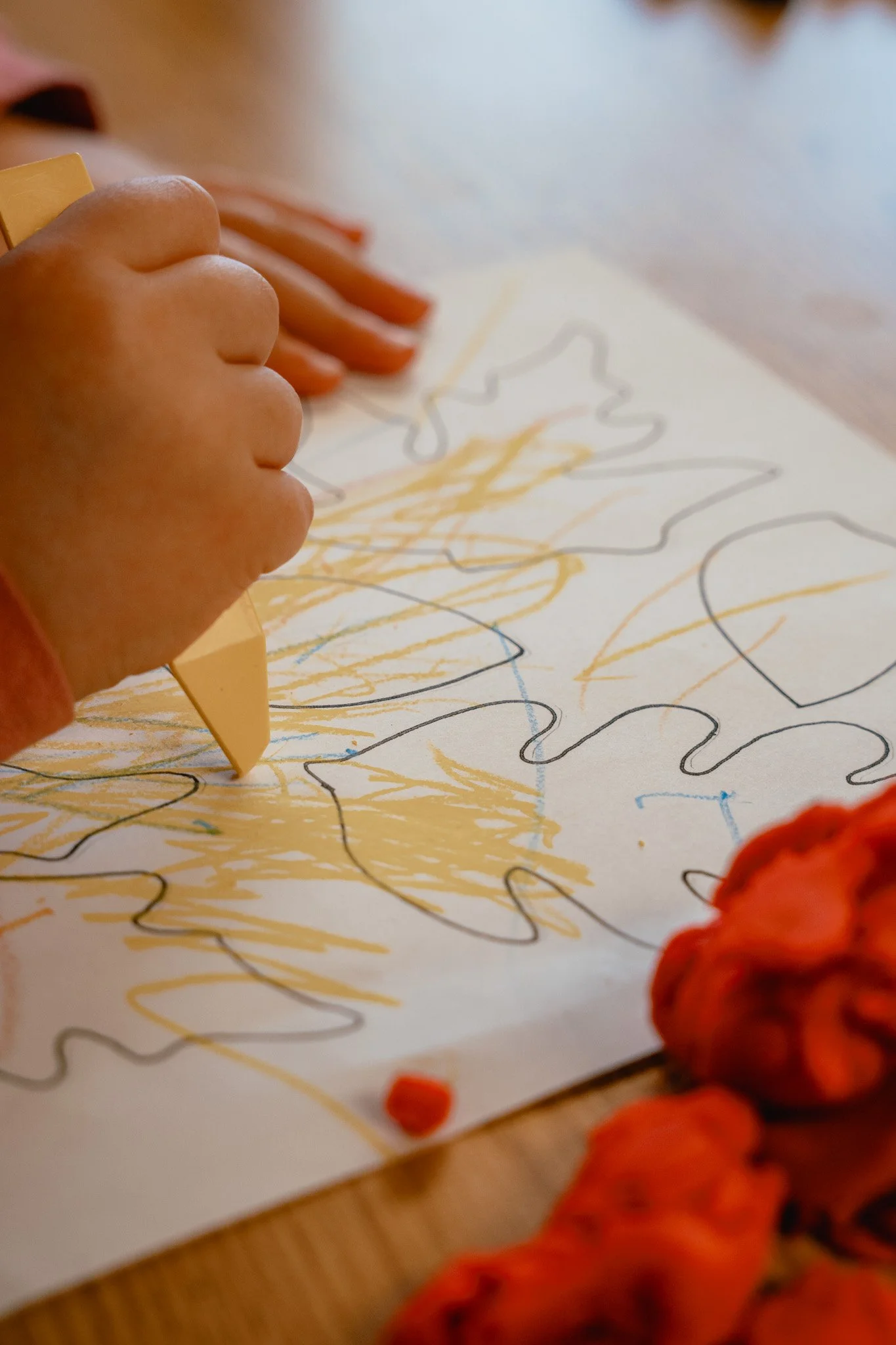 Child's hand coloring a large paper with a yellow crayon, creating a scribbled and outlined picture. There are orange objects, possibly made of textured material, near the paper.