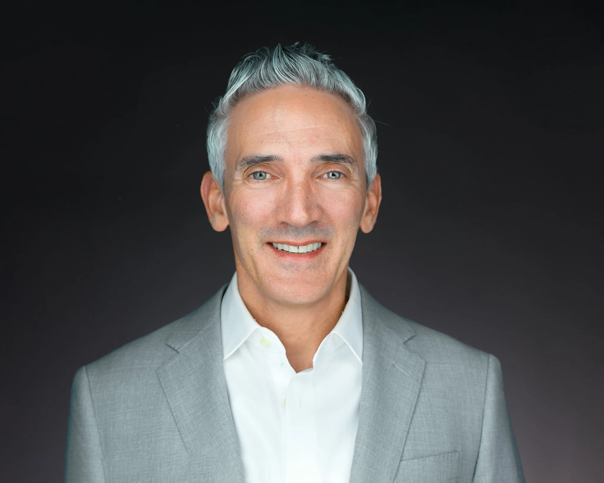 A middle-aged man with gray hair, wearing a light gray suit and white dress shirt, smiling against a dark background.