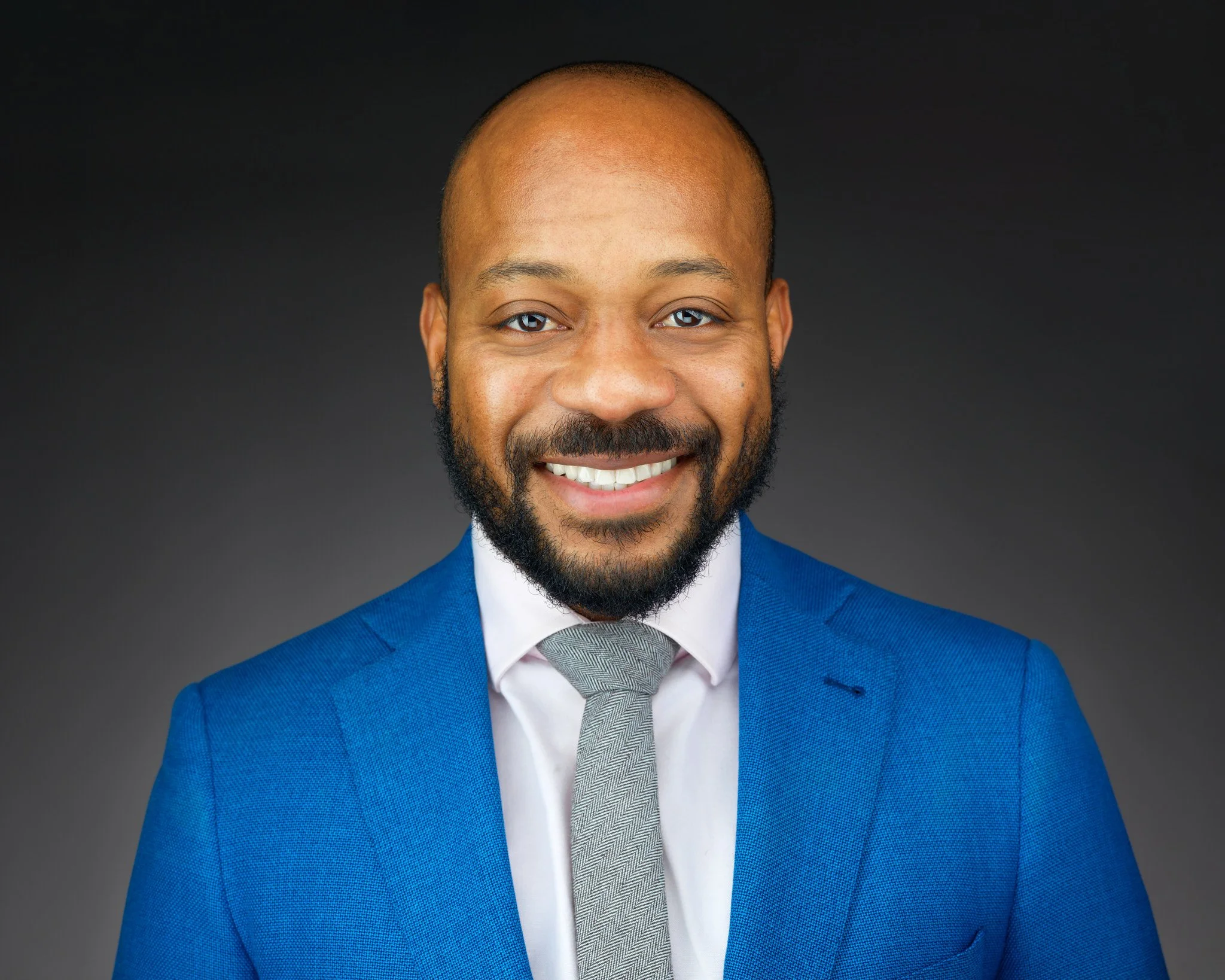 Headshot of a man with a beard and mustache wearing a blue suit, white shirt, and gray tie, smiling against a dark gray background.
