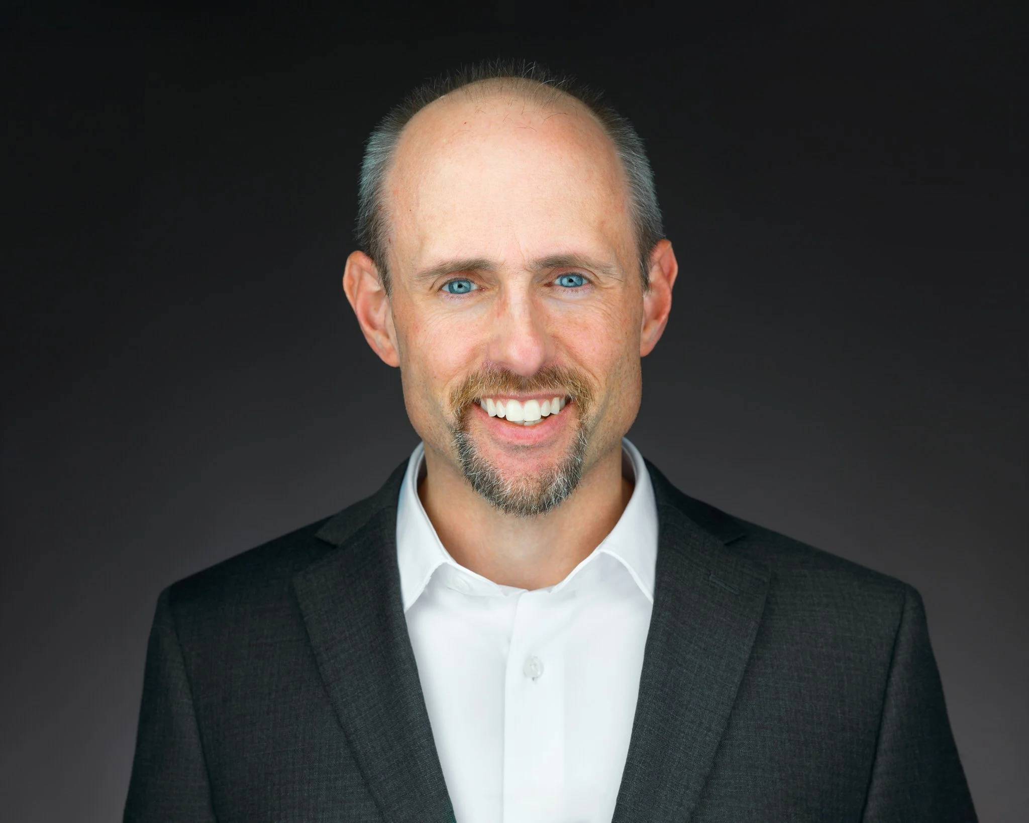 Portrait of a man with light skin, blue eyes, short gray hair, a mustache and goatee, wearing a black suit jacket and a white shirt against a dark background.