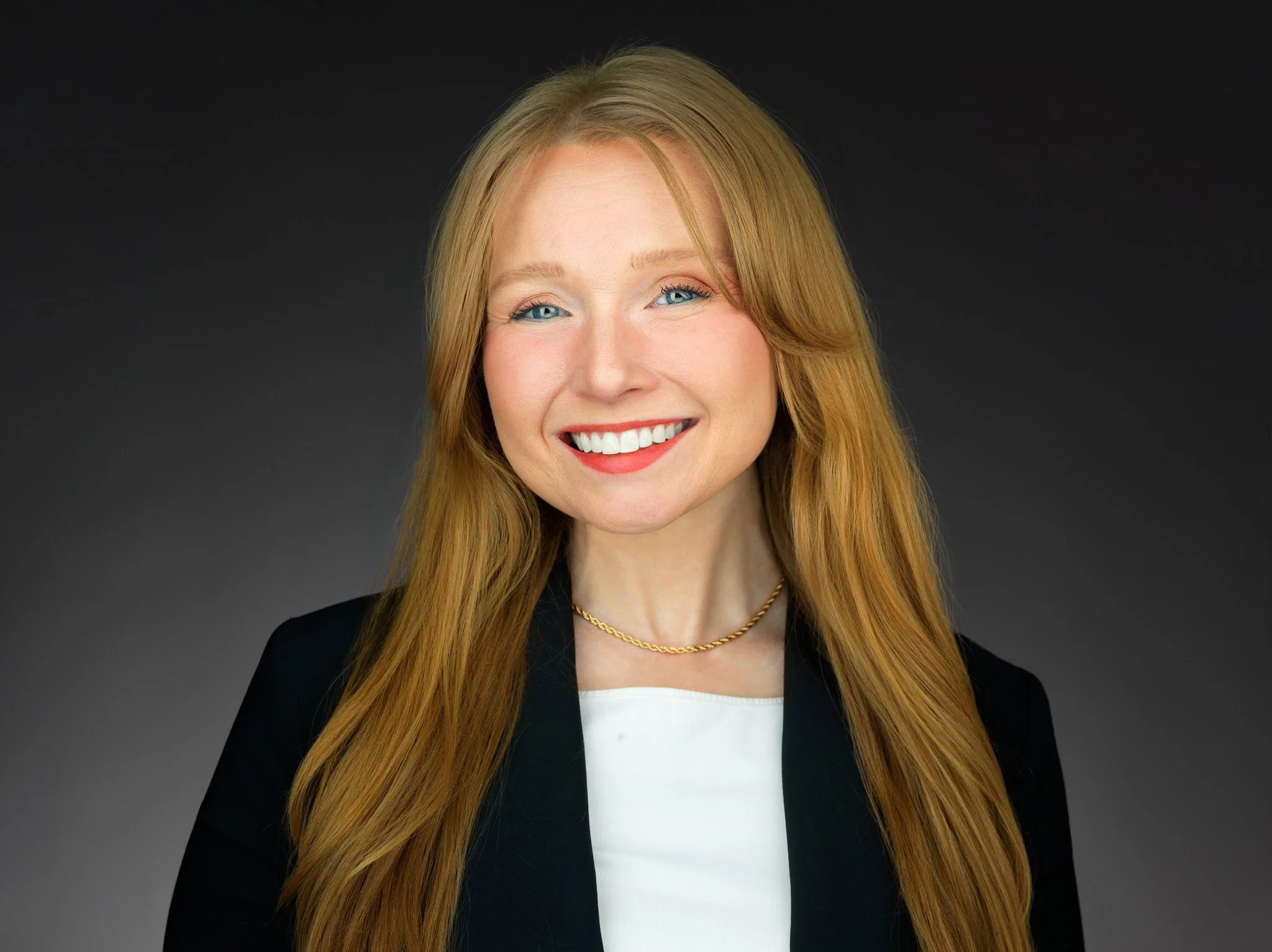Professional headshot of a smiling woman with long red hair, wearing a black blazer, white blouse, and a gold necklace, against a dark background.