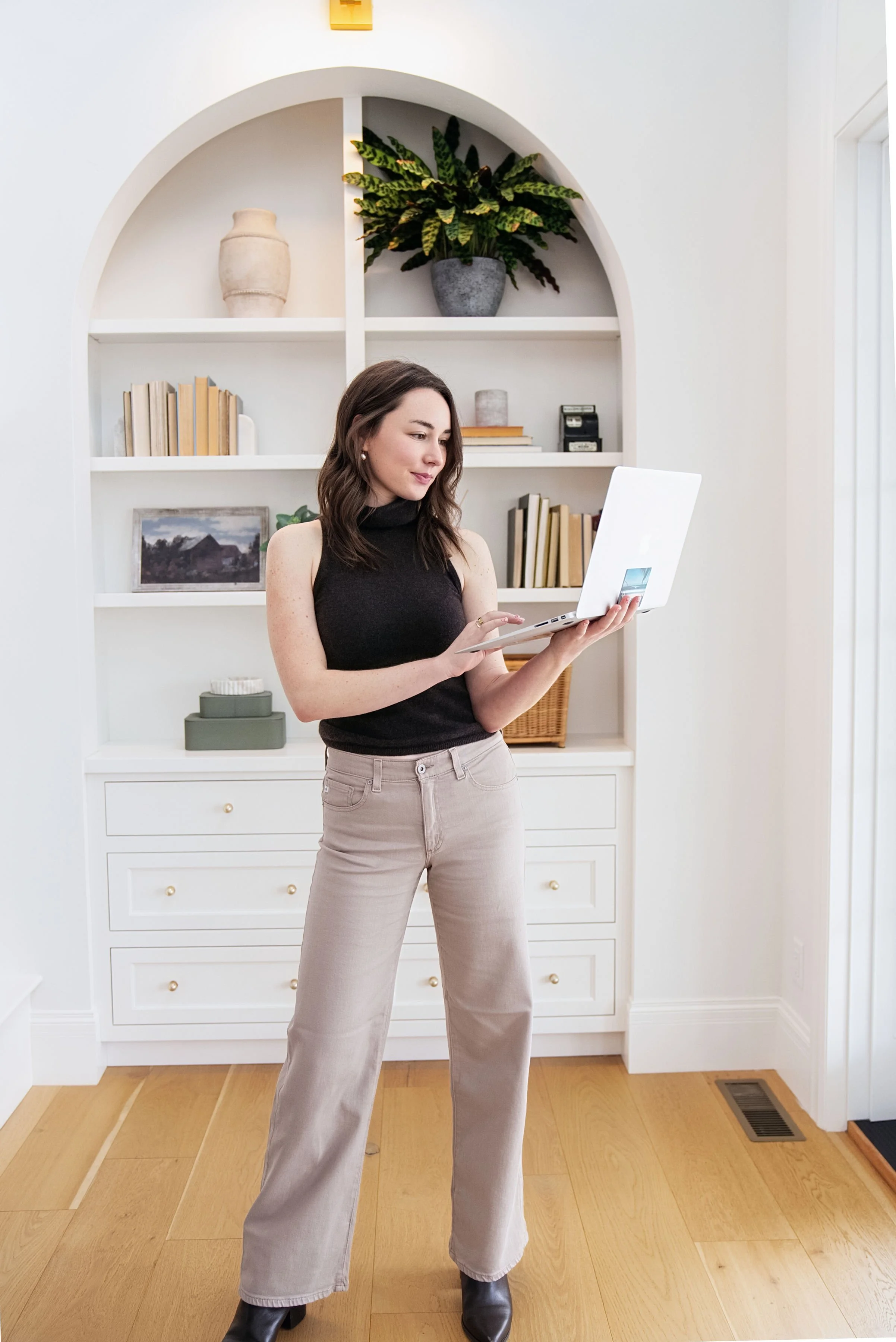 A young woman with shoulder-length dark brown hair, wearing a black sleeveless turtleneck and beige wide-leg trousers, standing in a bright room with hardwood floors. She is holding a white laptop and looking at it, with a bookshelf with books and decor items behind her.