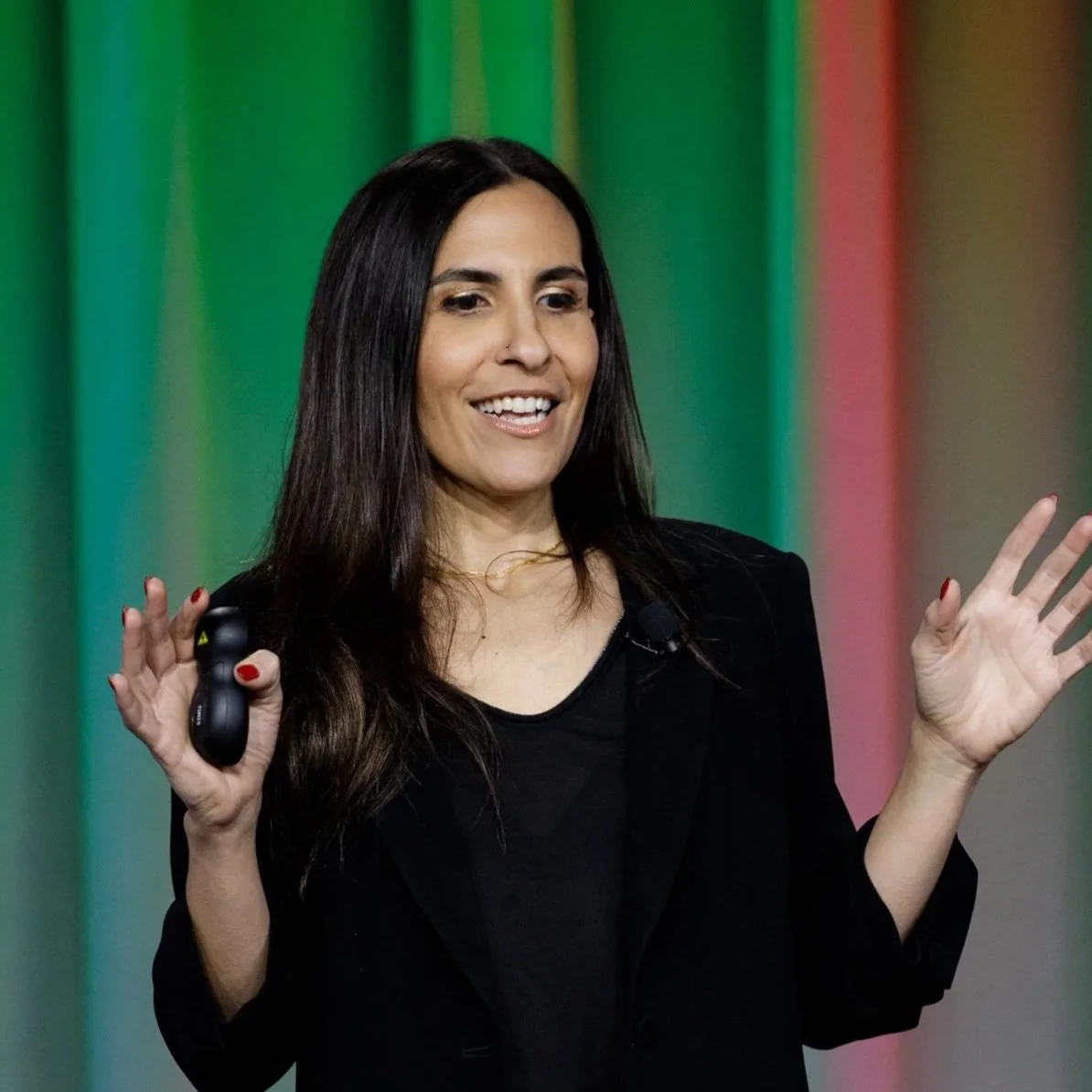 A woman with long dark hair, wearing a black blazer and a black shirt, smiling, holding a presentation remote, gesturing with her right hand during a speech or presentation against a colorful green and red background.