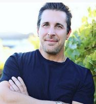 A man with dark hair and a beard, wearing a navy blue shirt, standing outdoors with greenery behind him.
