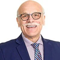 Photograph of an elderly man with glasses, dressed in a blue suit and striped shirt, smiling against a neutral background.