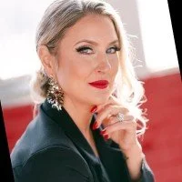 A woman with blonde hair and red lipstick, wearing a black blazer, sitting in front of a window with a cityscape background.