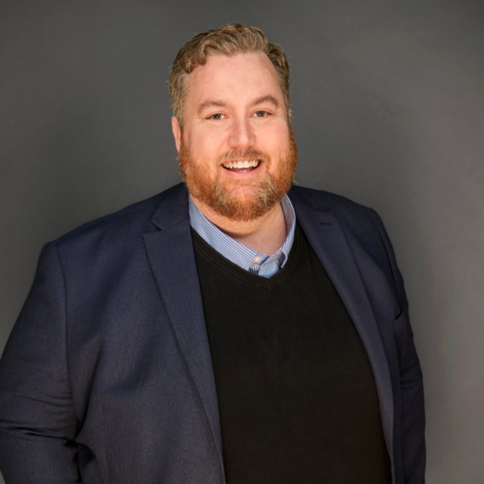 A smiling man with a beard wearing a navy blazer, light blue striped dress shirt, and black sweater against a gray background.