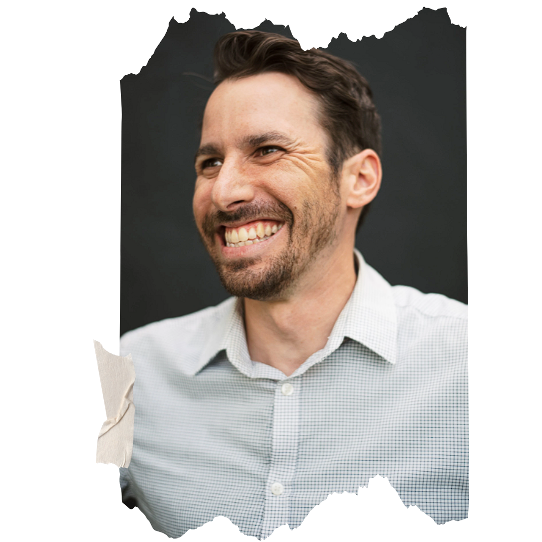 A smiling man with short dark hair, a beard, wearing a light gray button-up shirt, against a black background.