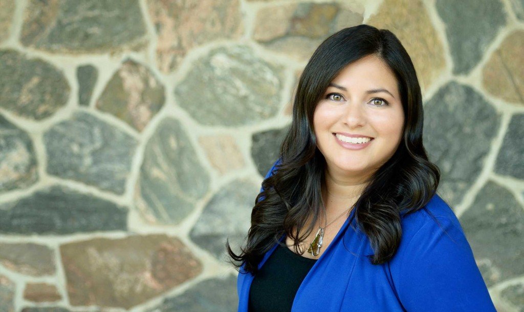A smiling woman with long dark hair wearing a blue jacket and a black top, standing in front of a stone wall.