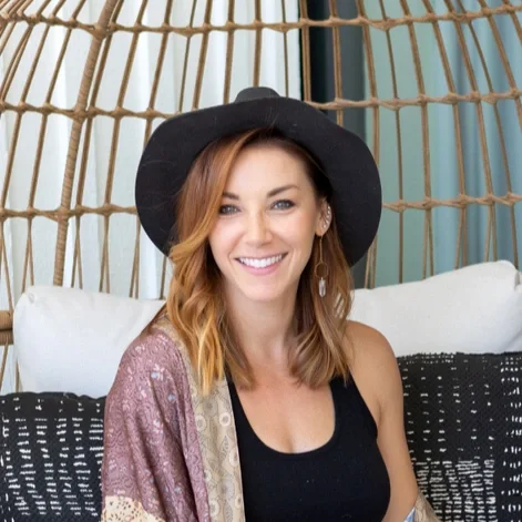 A smiling woman with shoulder-length red hair wearing a black hat and black tank top, sitting on a white cushion in a wicker hanging chair.