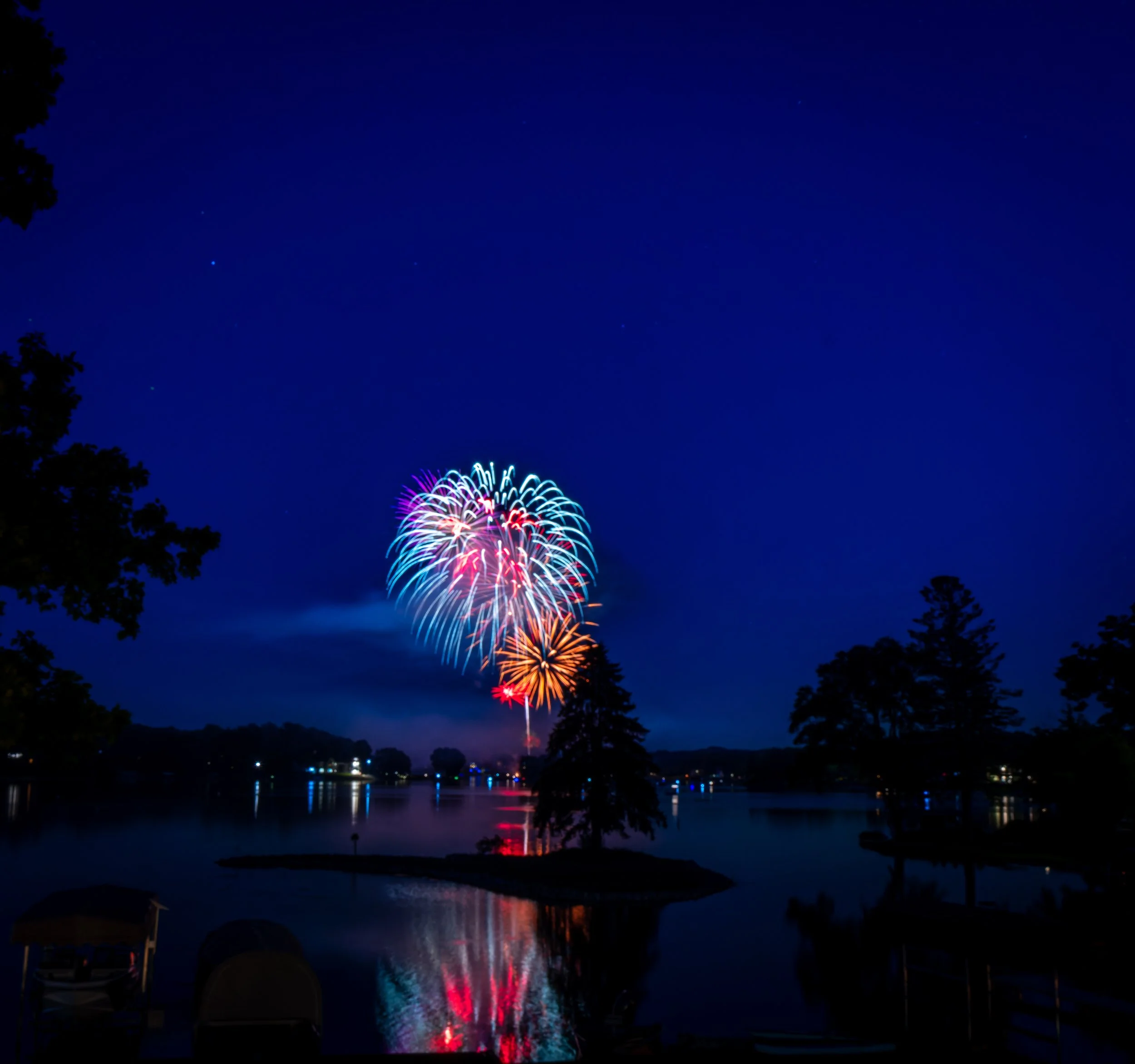 Colorful fireworks display over a lake at night, with reflections on the water and silhouetted trees in the foreground.