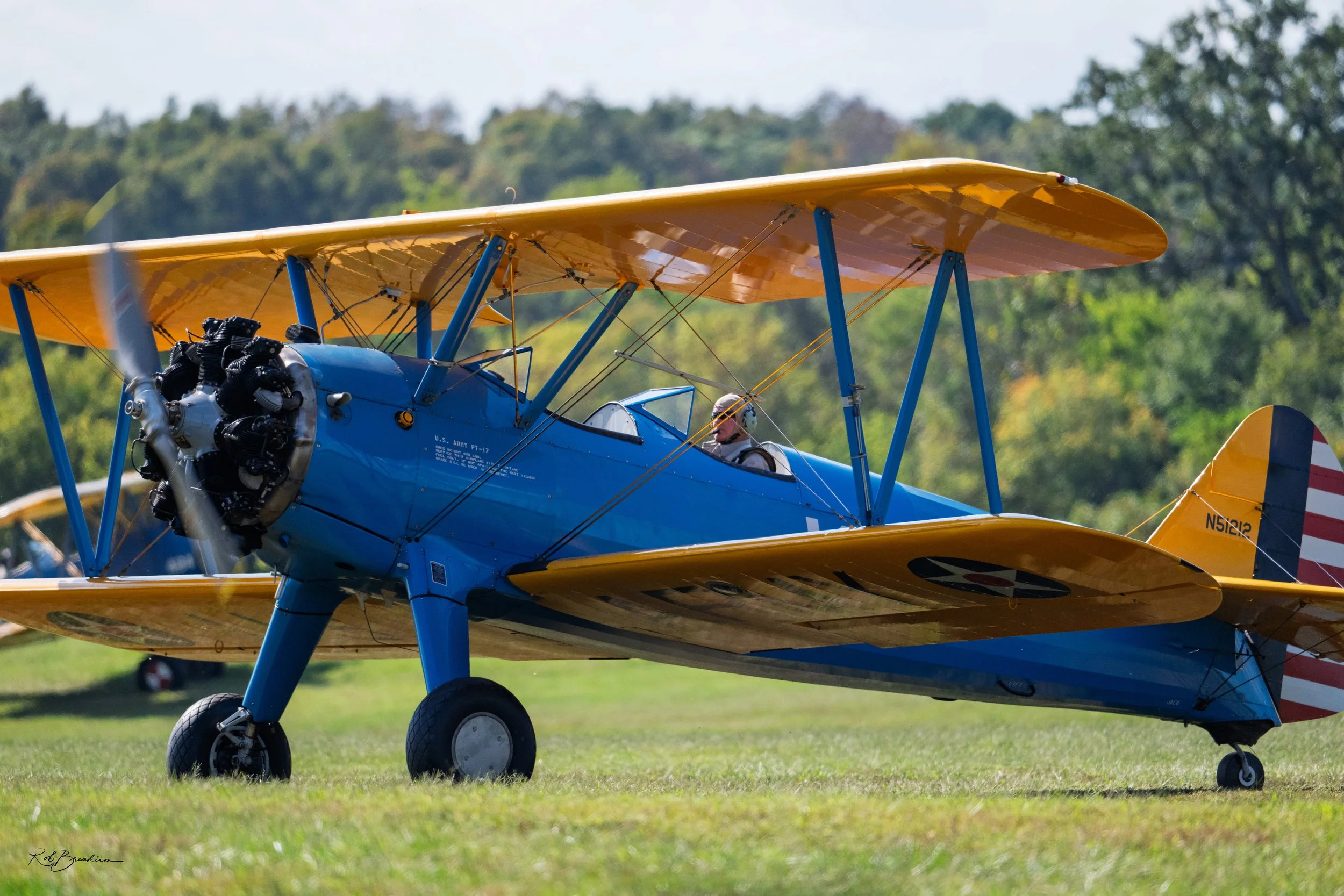 A vintage U.S. Army biplane with a blue fuselage and yellow wings, parked on a grassy field with trees in the background.