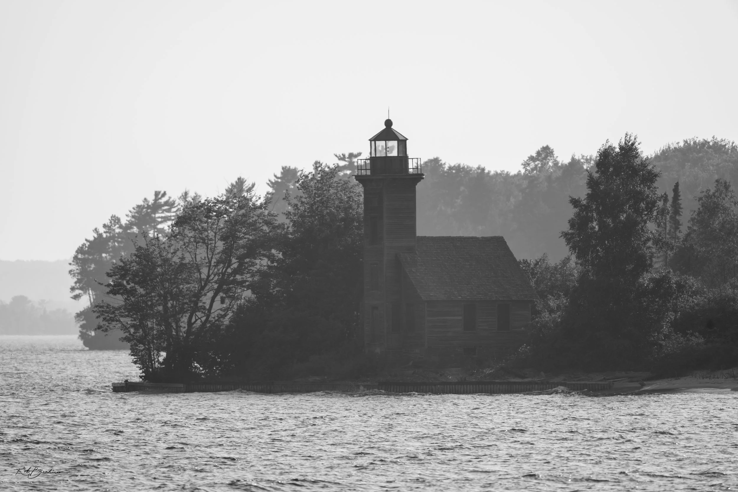 Black and white photo of a seaside lighthouse on a small island, surrounded by trees, with water in the foreground and a faint outline of land in the background.