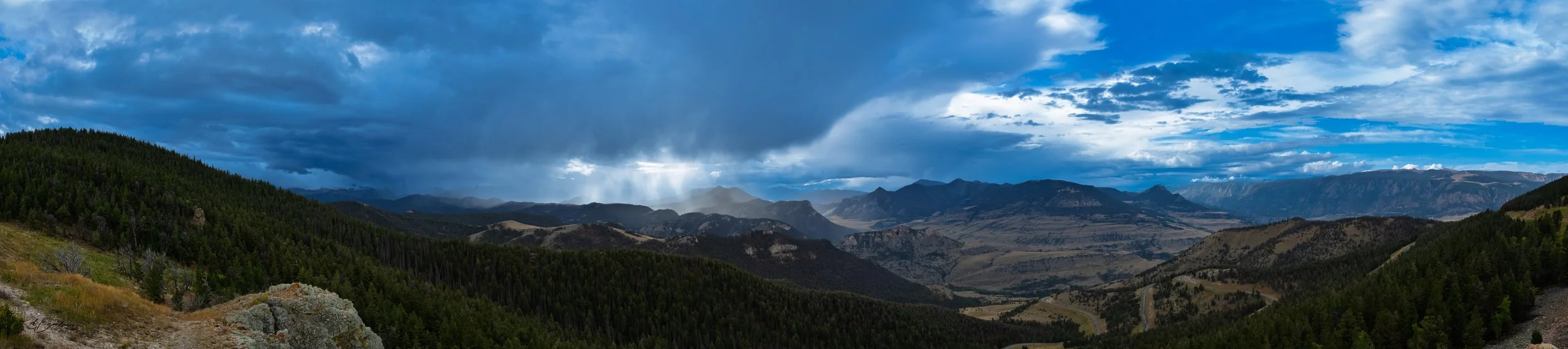Panoramic view of a mountainous landscape under a partly cloudy blue sky with rain in the distance.