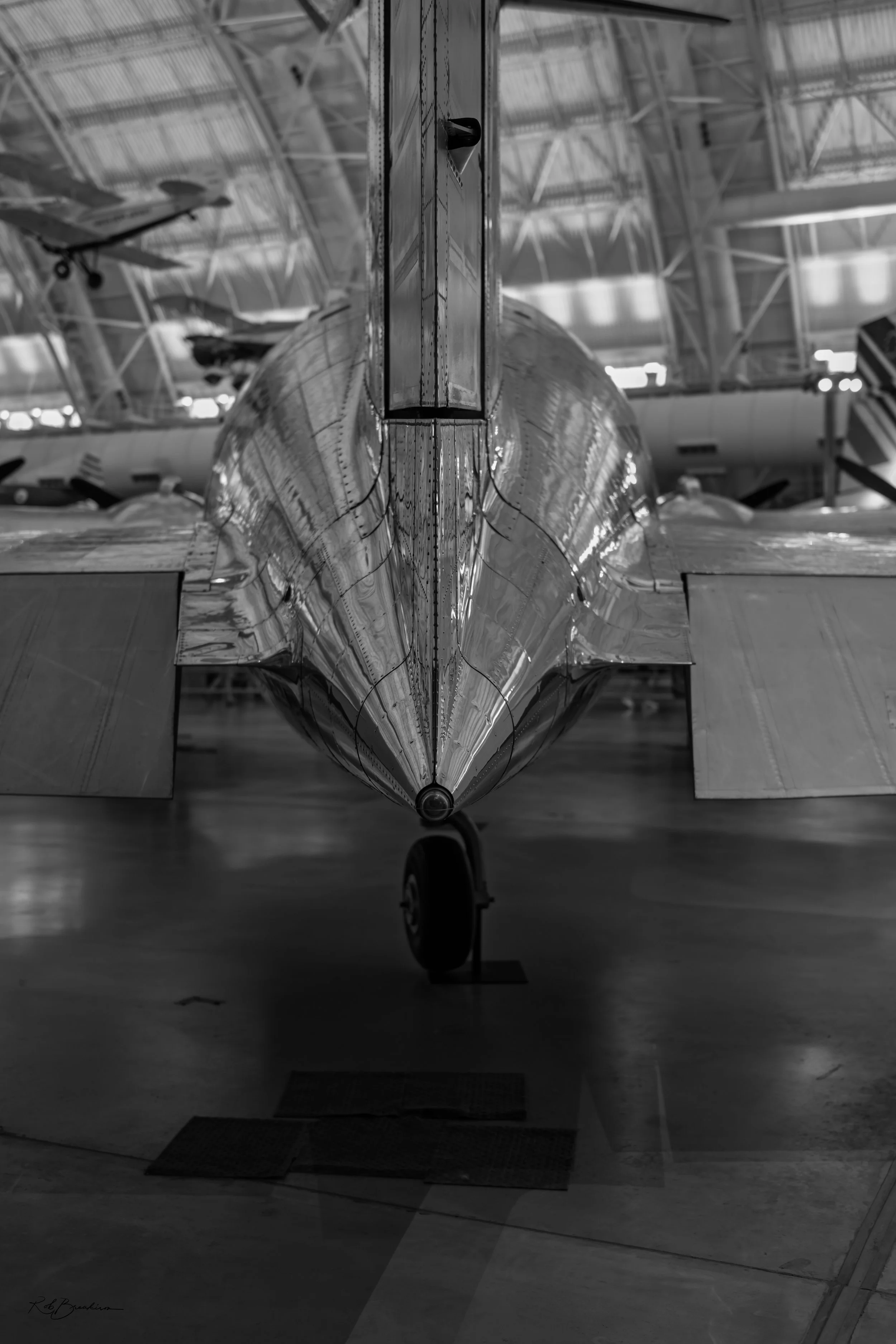 Black and white photo of a commercial airplane inside an aircraft hangar, viewed from the front showing the nose cone and front landing gear, with other aircraft and the hangar's roof structure in the background.