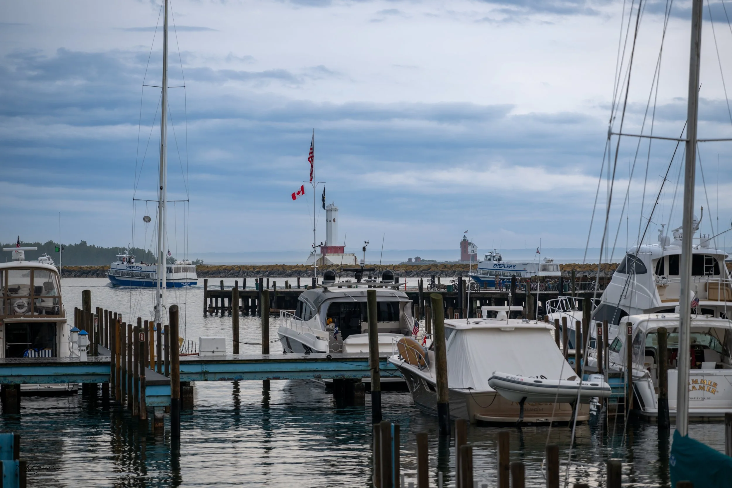 A marina with several boats docked, including sailboats and motorboats, with a breakwater and two lighthouses in the background under an overcast sky.