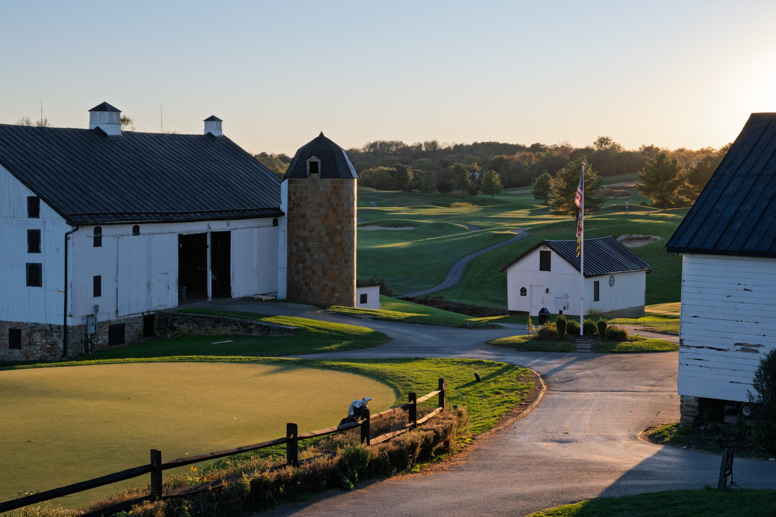 A scenic view of a golf course with a white barn-style building, a smaller white structure with a flag, a cart path, and lush green landscape with trees in the background at sunset.