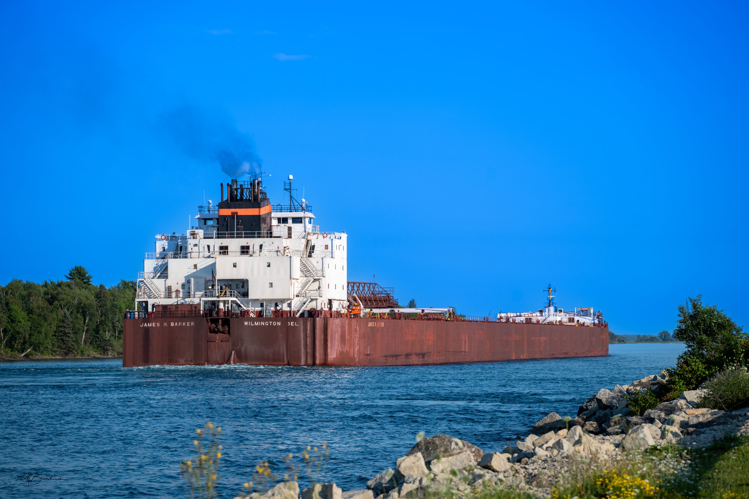 A large cargo ship named James R. Barker sailing on a river with a rocky shoreline and green trees, under a clear blue sky.