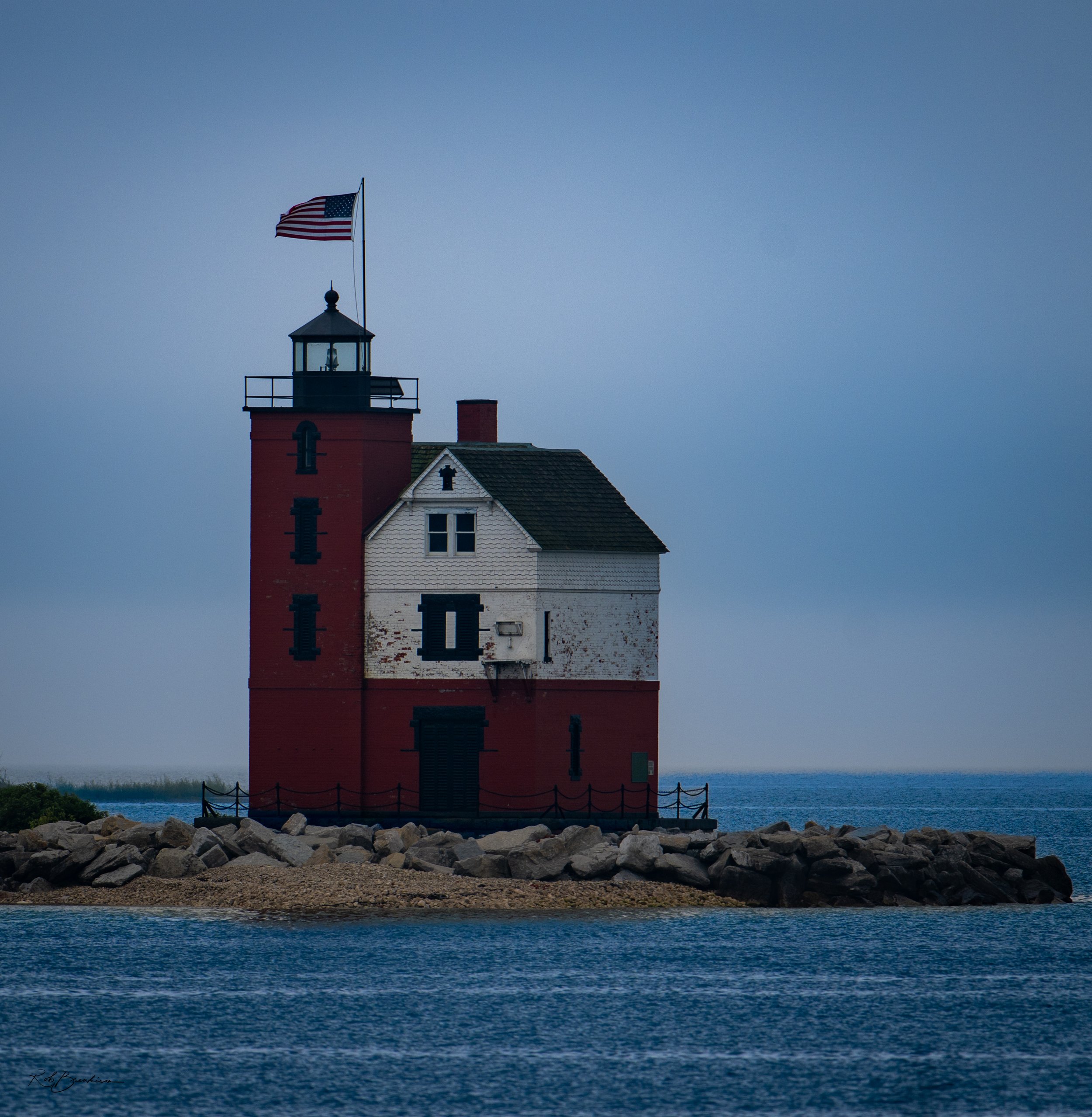 A lighthouse with a red base, white upper structure, and black roof, situated on a rocky breakwater in the ocean. An American flag flies atop the lighthouse against a cloudy sky.