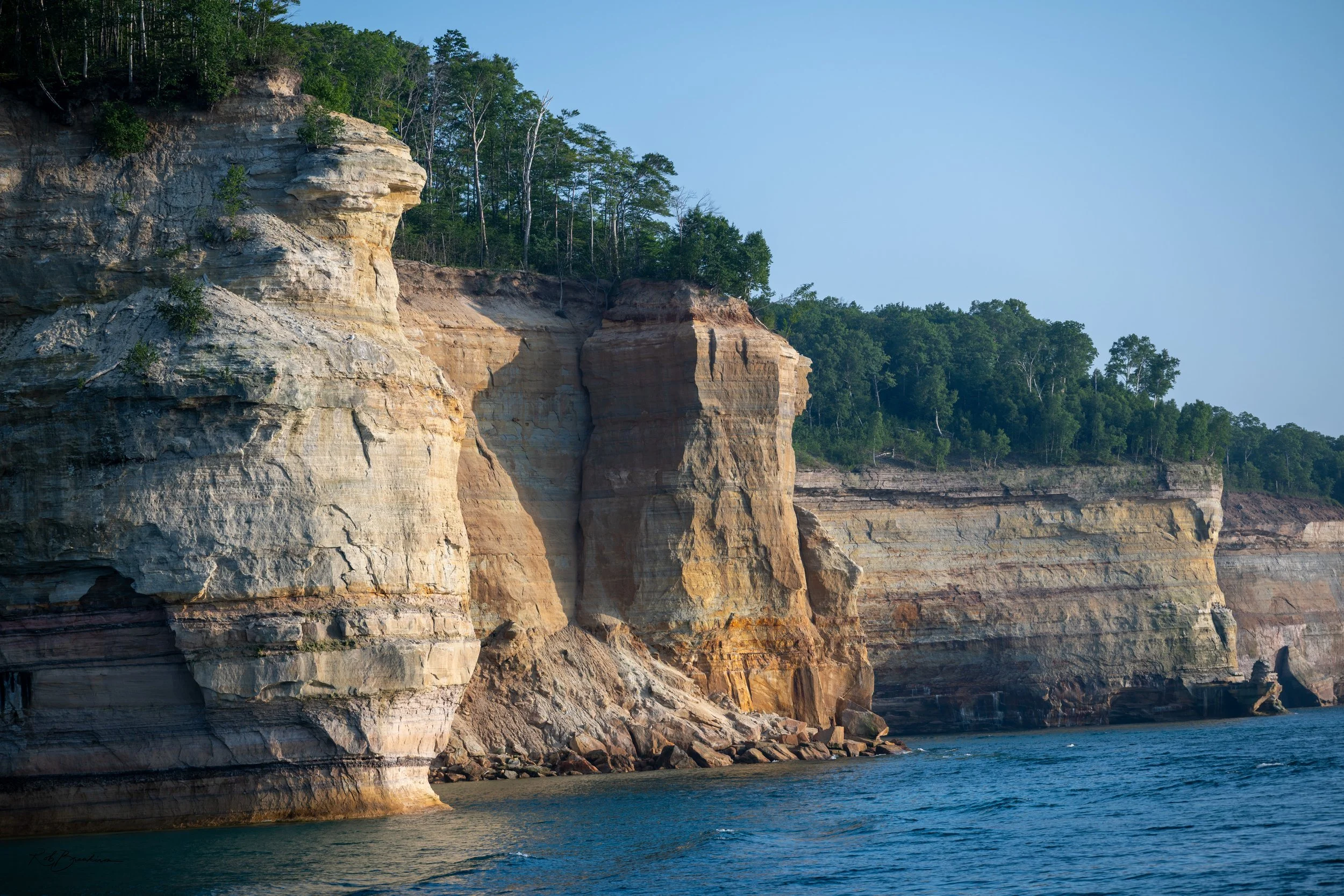 Cliffs along a body of water with trees at the top under a clear blue sky.