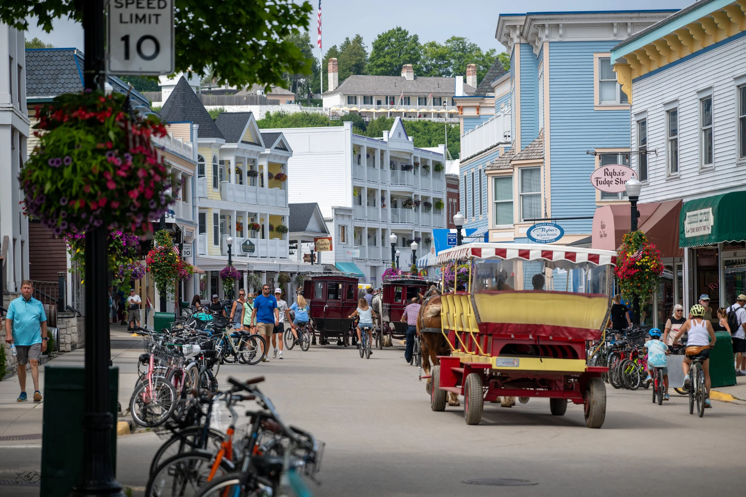 A busy street with people riding bicycles, walking, and a horse-drawn carriage. Colorful buildings line the street, with hanging flower baskets and storefront signs. There is a speed limit sign showing 10 miles per hour.