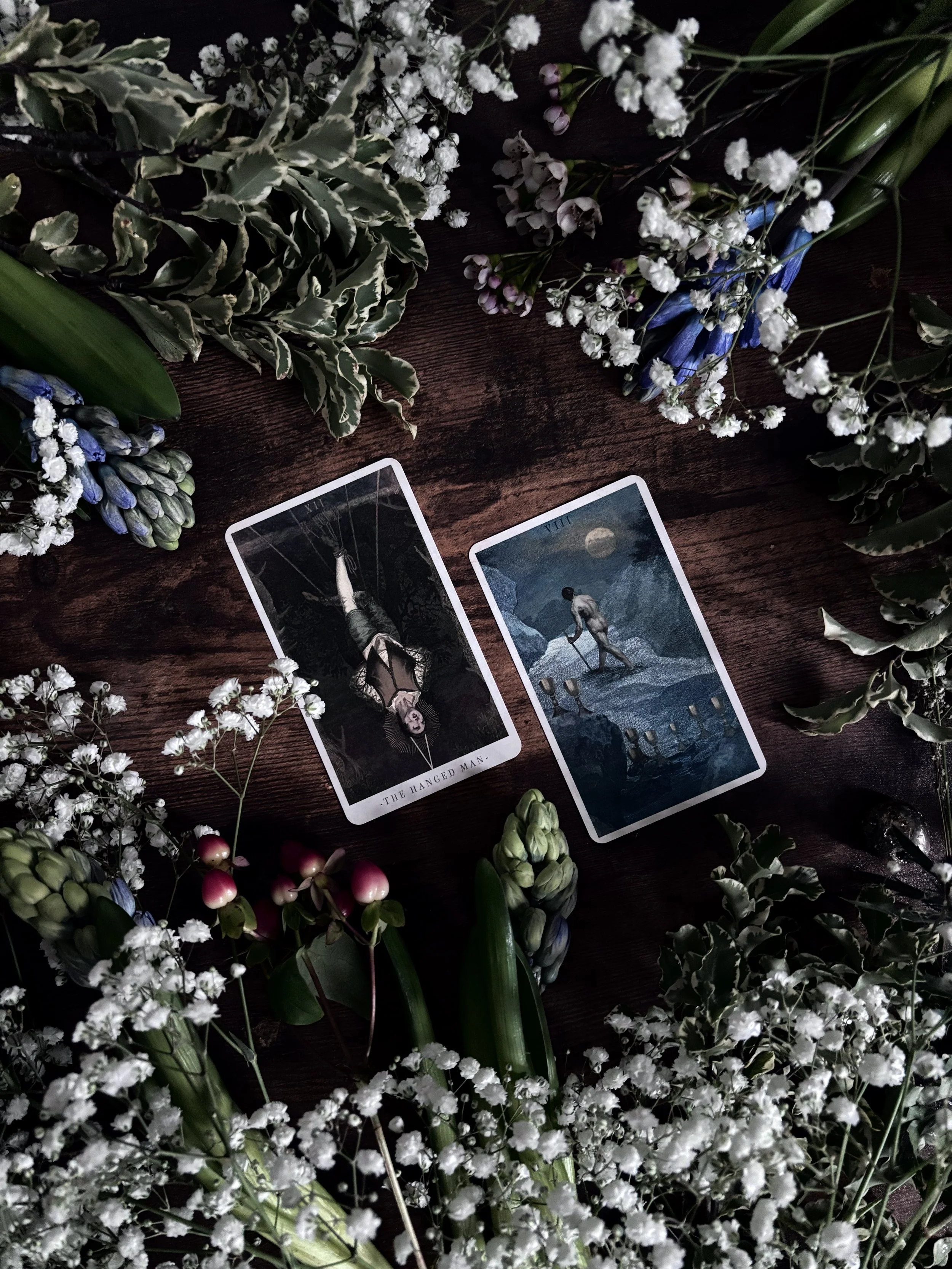 Tarot cards on a wooden surface surrounded by white baby's breath and variegated green plants. One card shows a woman hanging upside down, labeled "The Hanged Man." The other card depicts a figure by the moonlit water with mountains in the background.