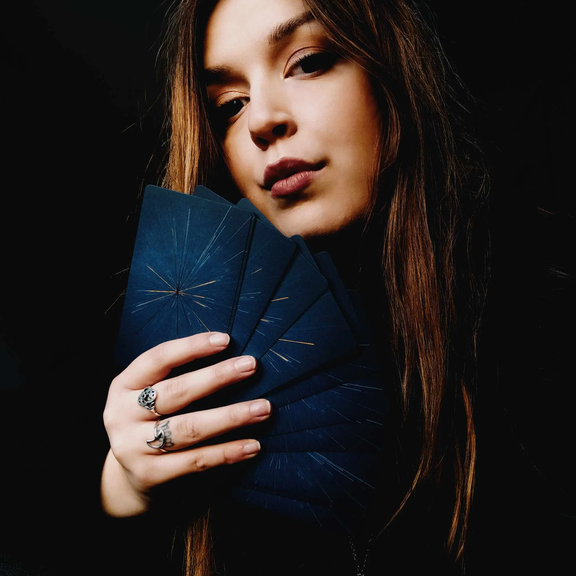 A young woman with long brown hair holding a fan with a dark blue and gold starburst design, wearing rings on her fingers, against a black background.