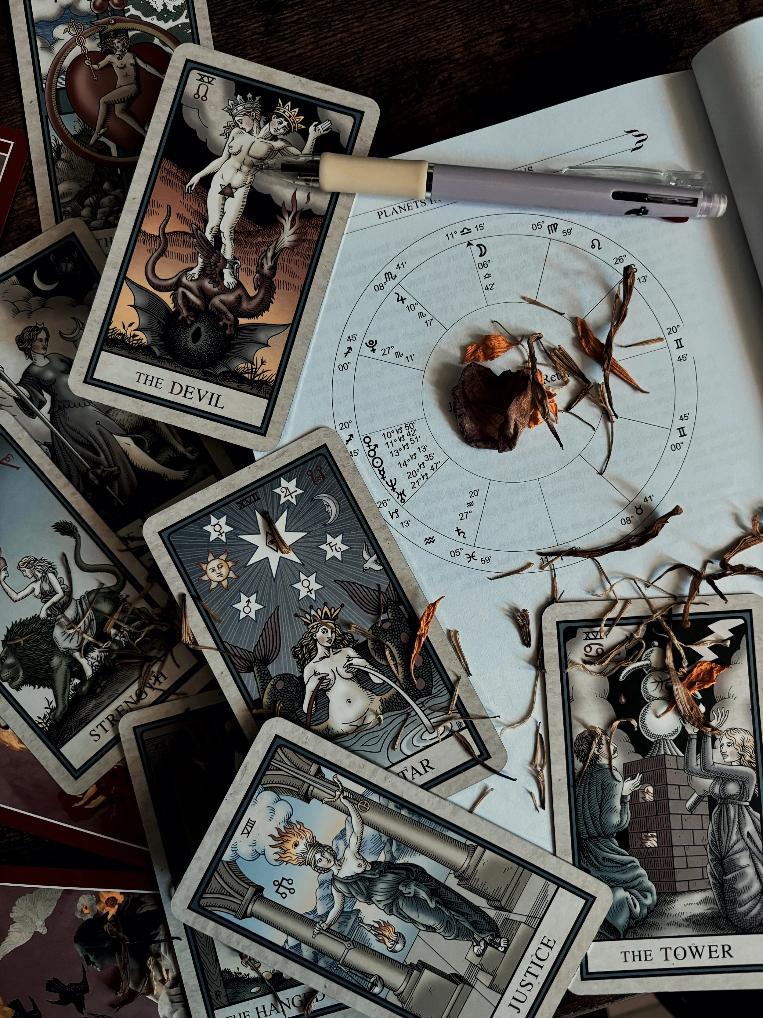 Tarot cards scattered on a table, including The Devil and Justice, dried flowers, a pen, and an astrology chart.