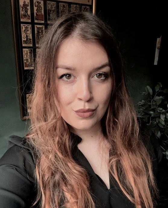 A young woman with long, wavy brown hair and light skin taking a selfie indoors, with framed artwork on the wall behind her and a dark green wall to her side.