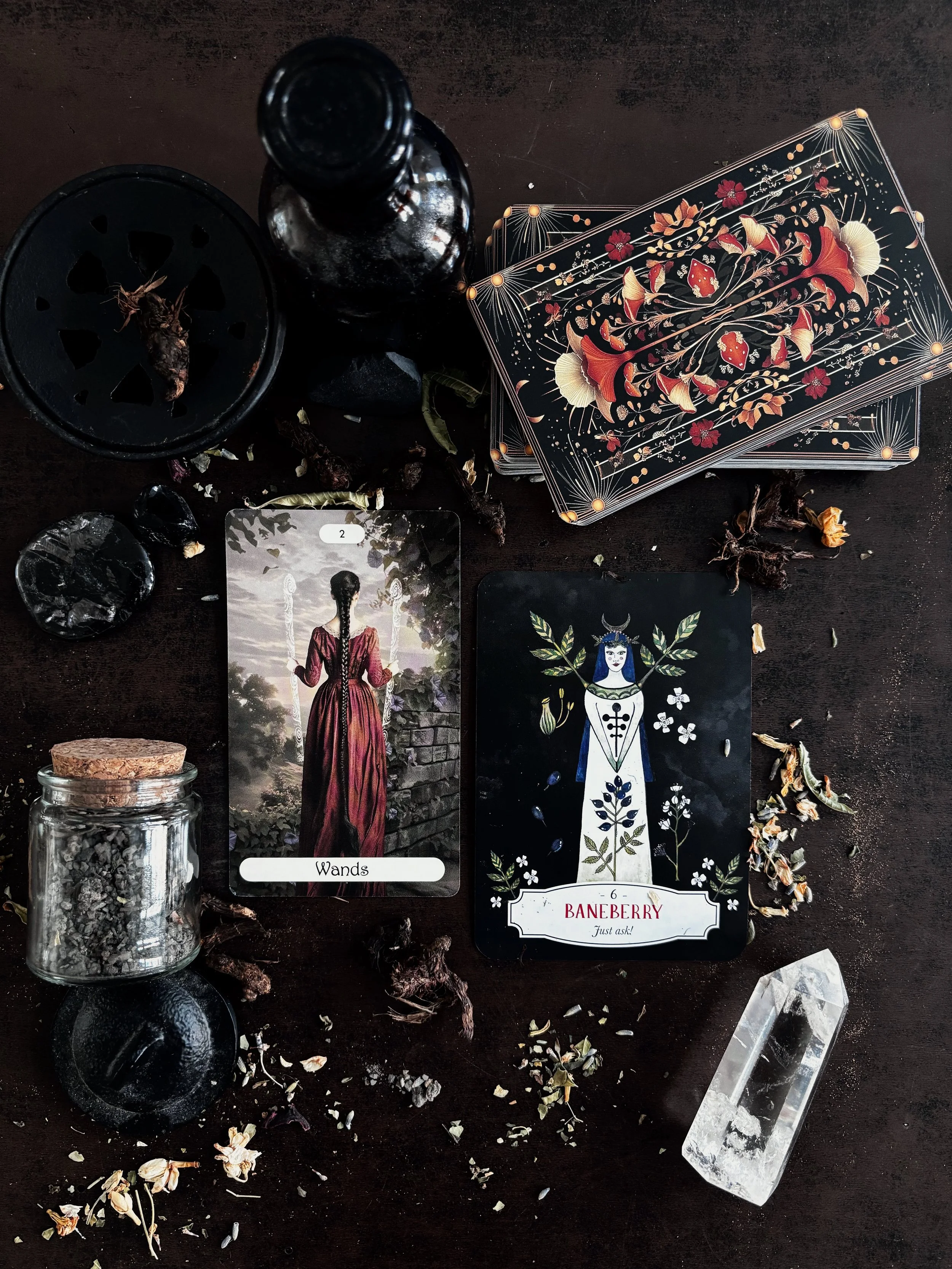 Tarot cards, crystals, a jar of herbs, and a black candle on a dark surface, used for divination or spiritual practices.