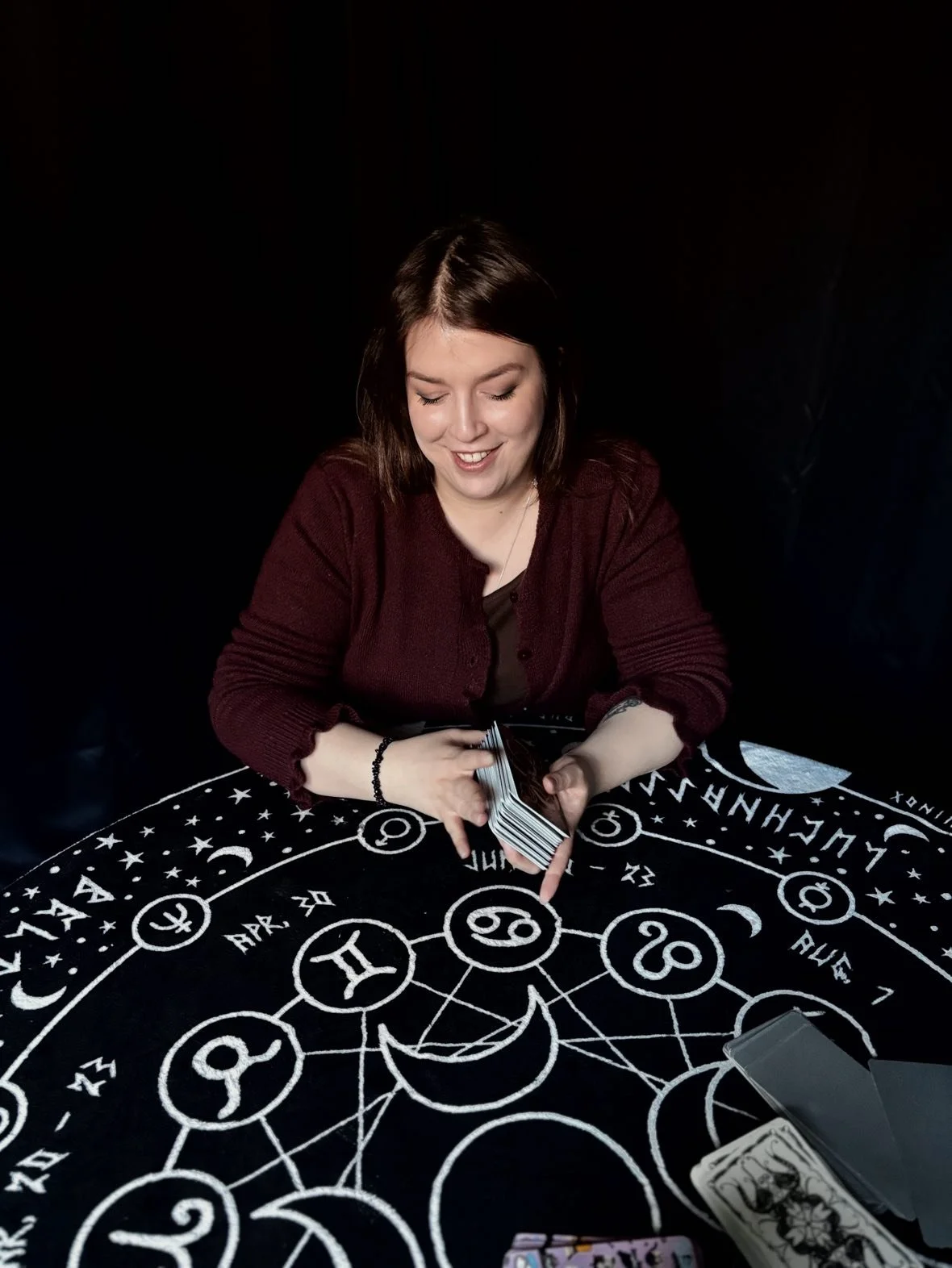 A woman with shoulder-length brown hair, wearing a maroon cardigan, smiles while holding tarot cards over a black cloth with white mystical symbols and zodiac signs.