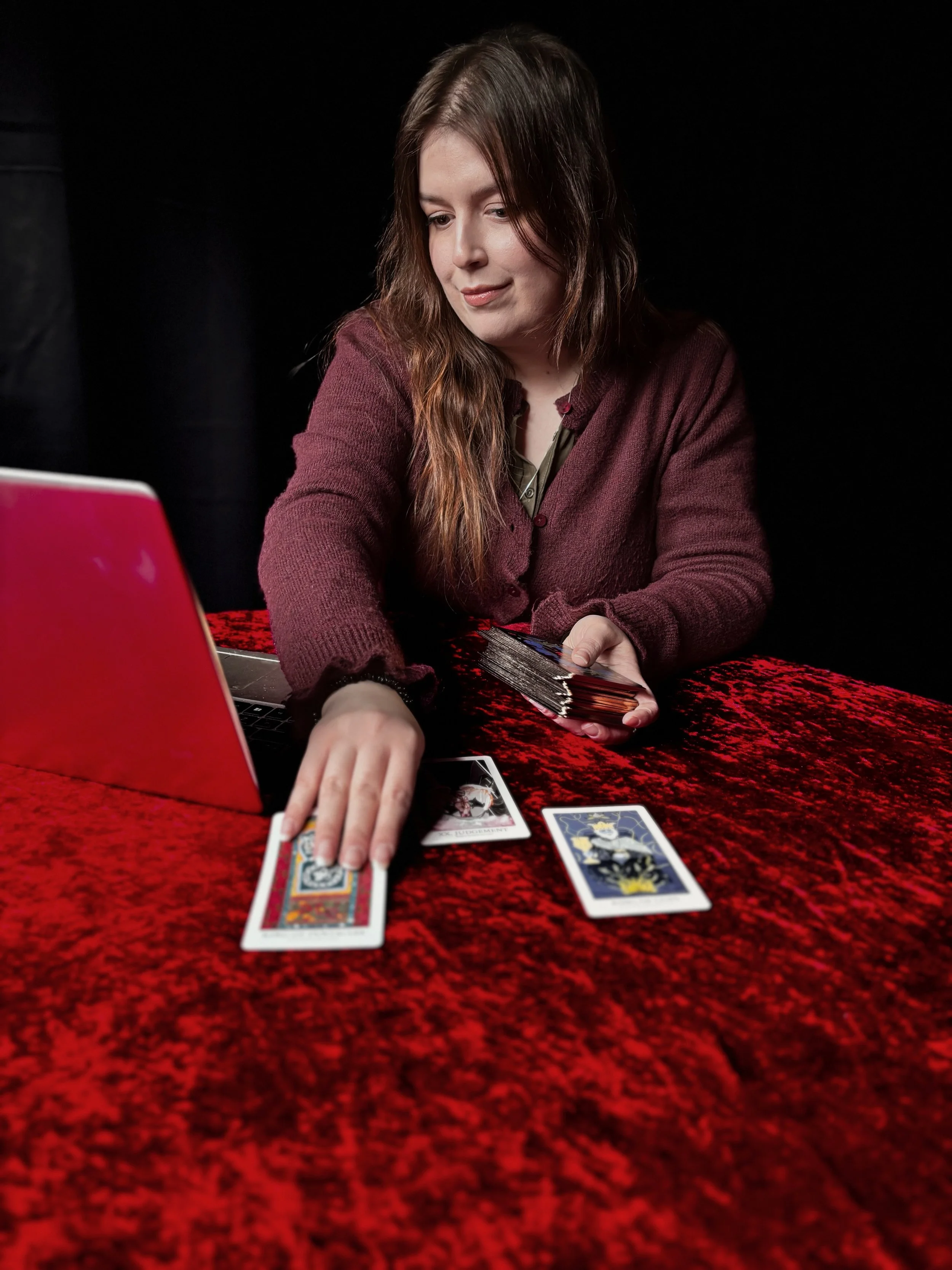 A woman with long brown hair wearing a maroon sweater sits at a red velvet table, reaching for tarot cards placed in front of her, with a laptop to her side against a black background.
