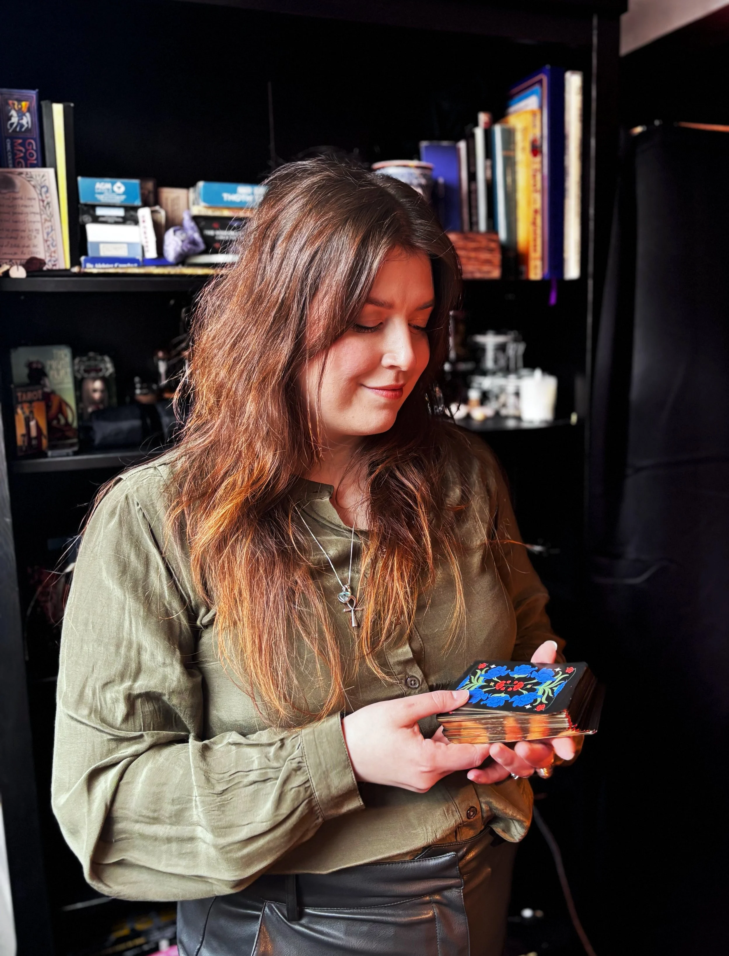 A young woman with long brown hair, wearing a green button-up shirt and a necklace with an ankh pendant, is standing indoors holding a deck of tarot cards with a floral design on the back, smiling softly as she looks down at the cards.