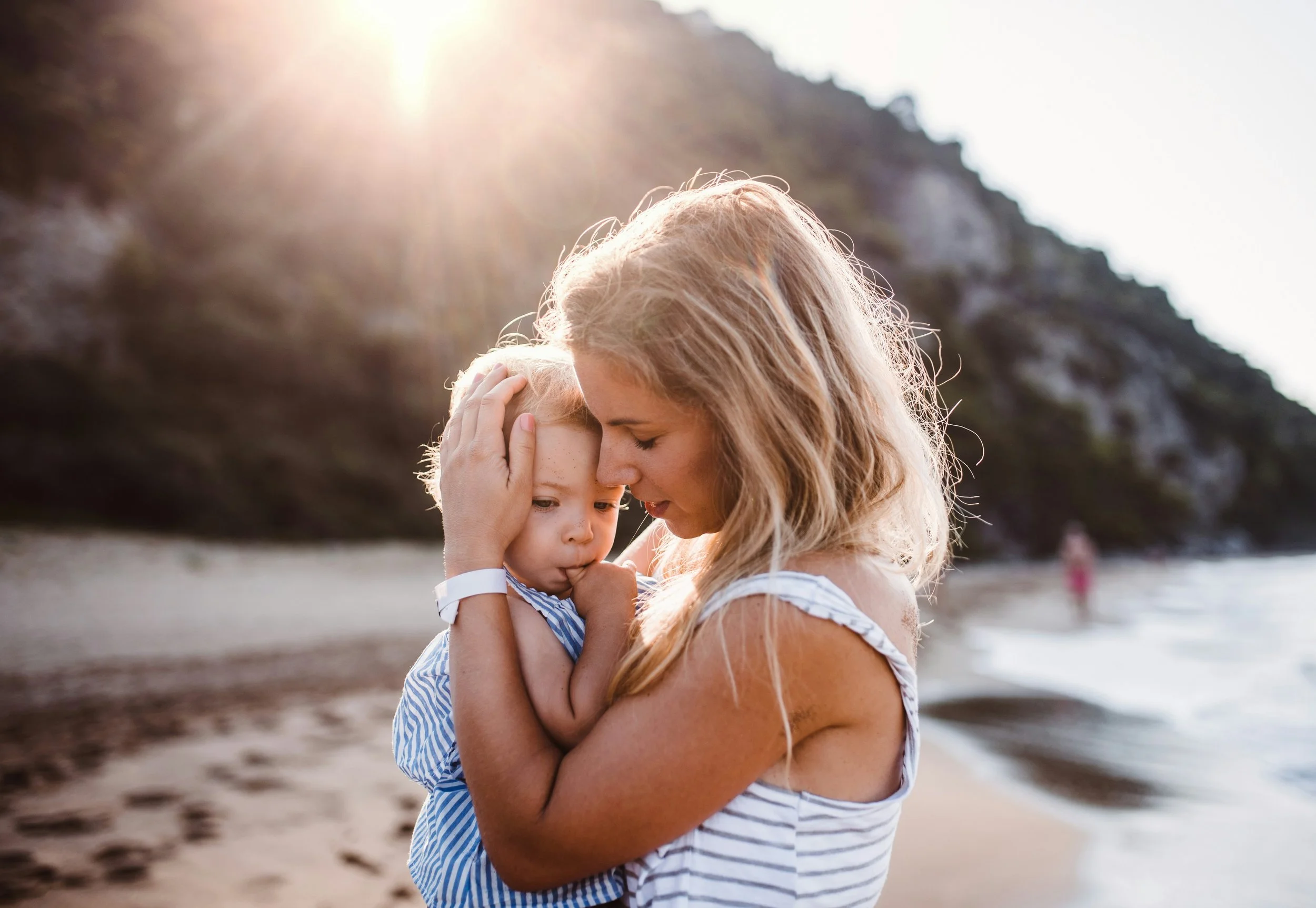 A woman holding a young child close to her face on a beach at sunset.