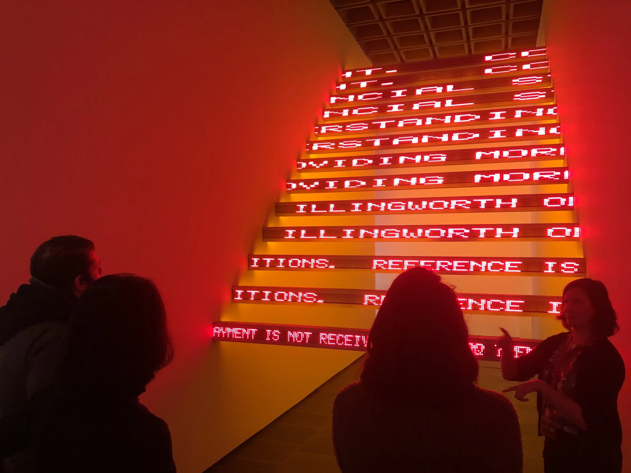 People standing in front of a Jenny Holzer installation with illuminated red LED signs displaying a series of words.
