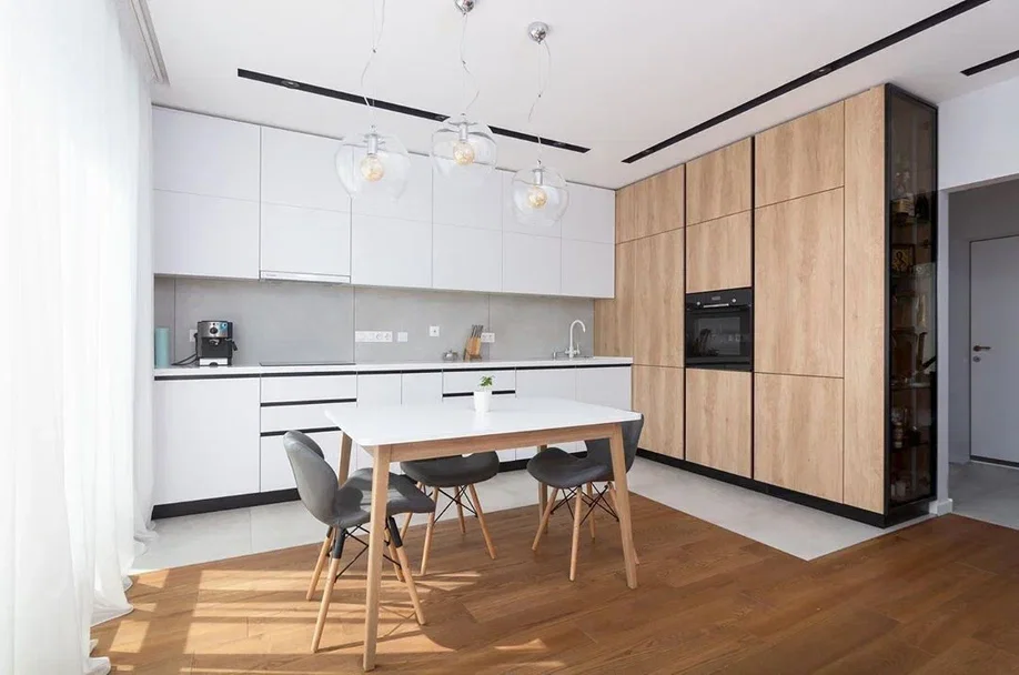 Modern kitchen with white upper and lower cabinets, natural wood panel cabinetry, a white dining table with four black chairs, pendant lights above, and a gray tiled backsplash.
