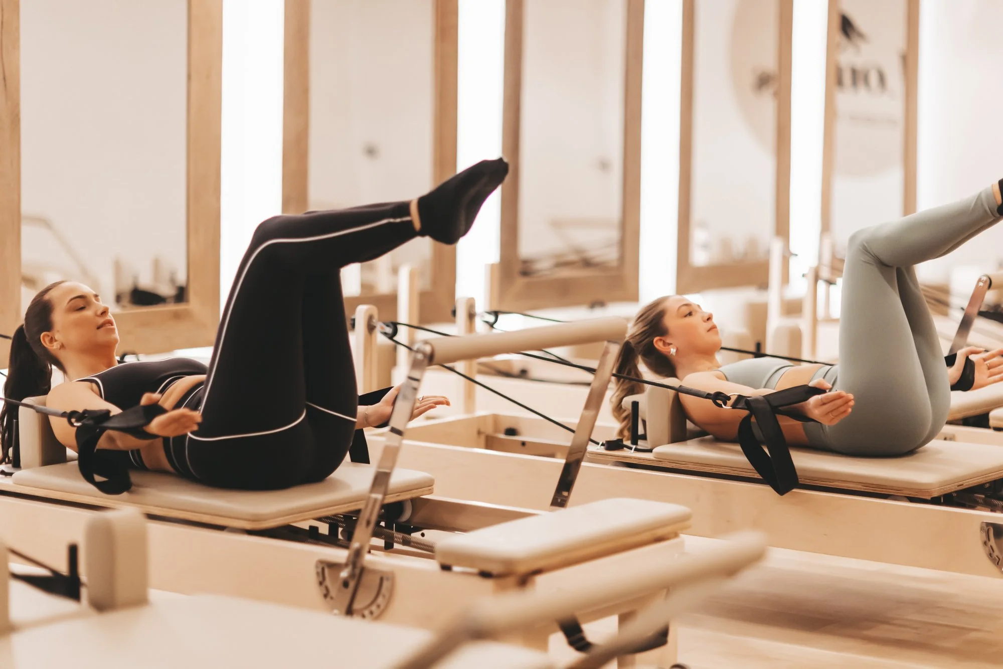 Deux femmes faisant de l'exercice sur des reformers Pilates dans un studio bien lumineux.