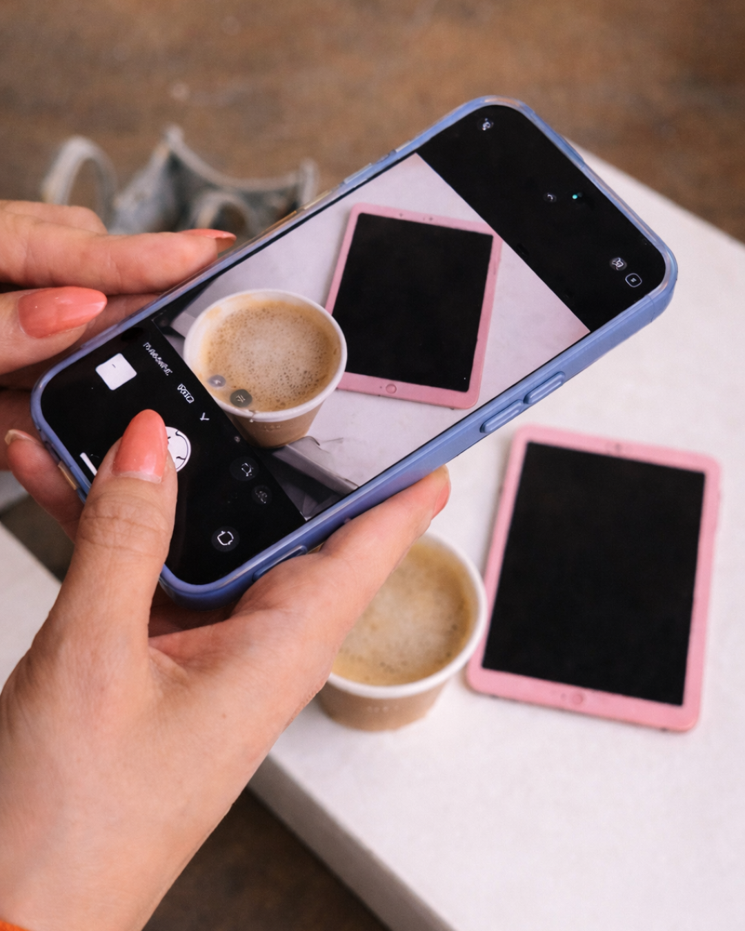 Person taking a photo of a cup of coffee with a smartphone, while two pink tablets are on a white table, and another cup of coffee is in the background.