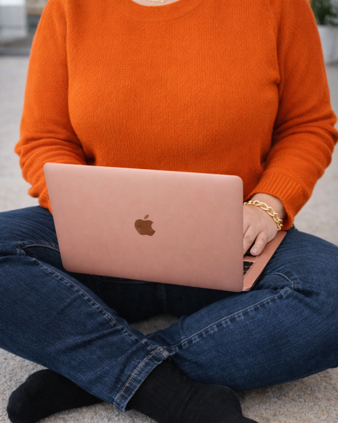 Person sitting cross-legged on the floor, wearing a bright orange sweater, using a rose gold MacBook laptop, with a gold bracelet on their wrist.