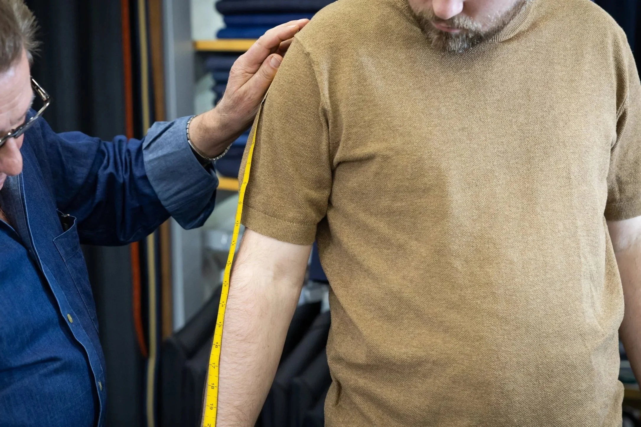 A person receiving a shoulder measurement with a tape measure by a professional in a fitting room or clothing store.