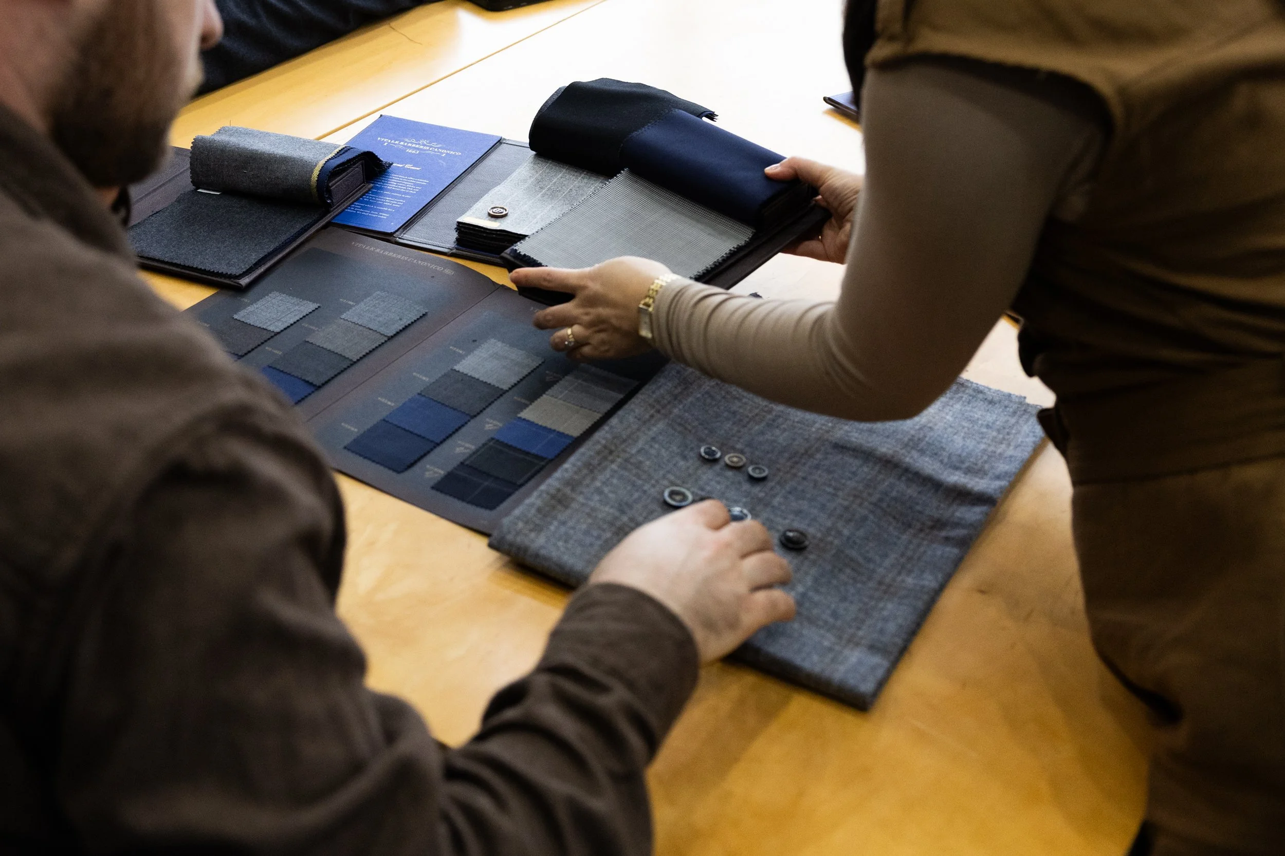 Two people are discussing fabric swatches and material samples on a wooden table, with fabric panels, buttons, and color samples.