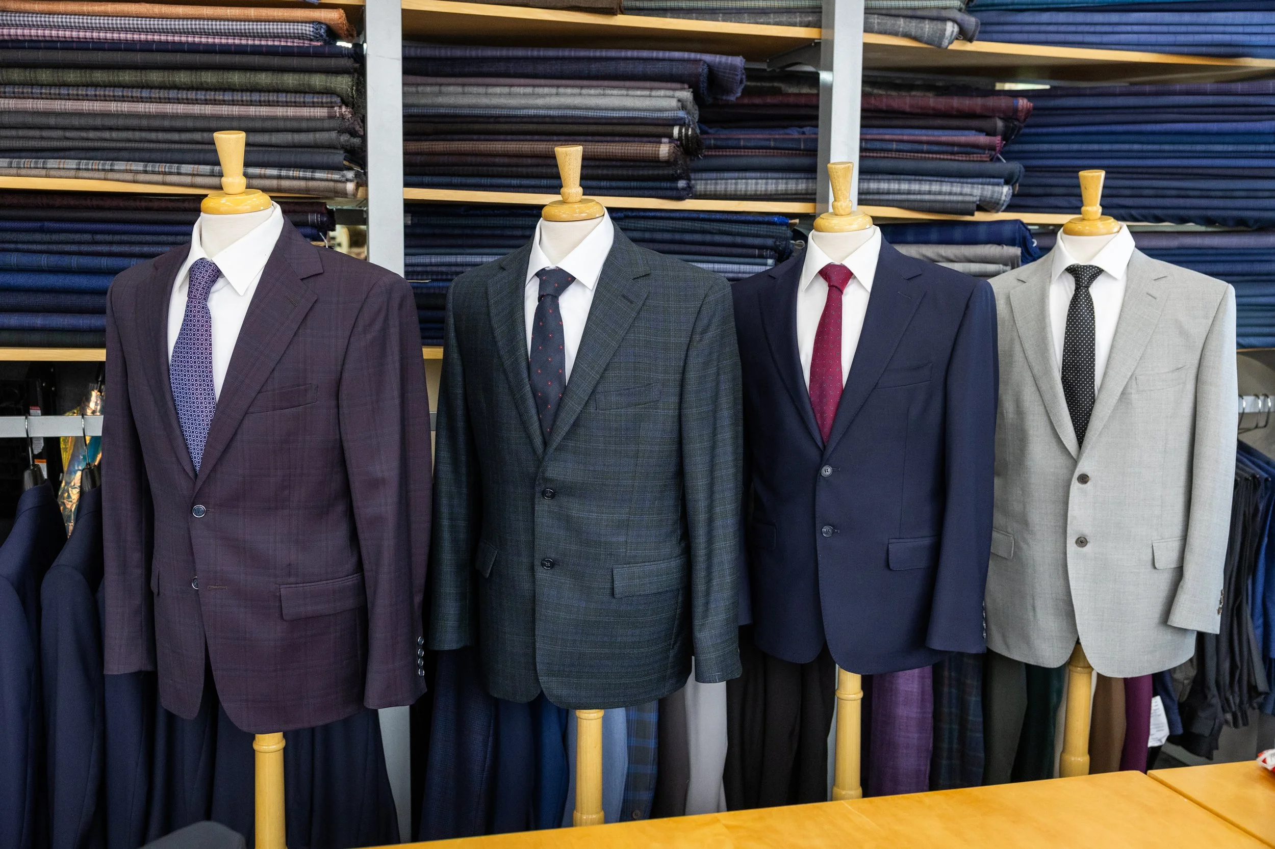 Display of four mannequins wearing business suits with shirts and ties, in front of shelves stacked with fabric bolts.