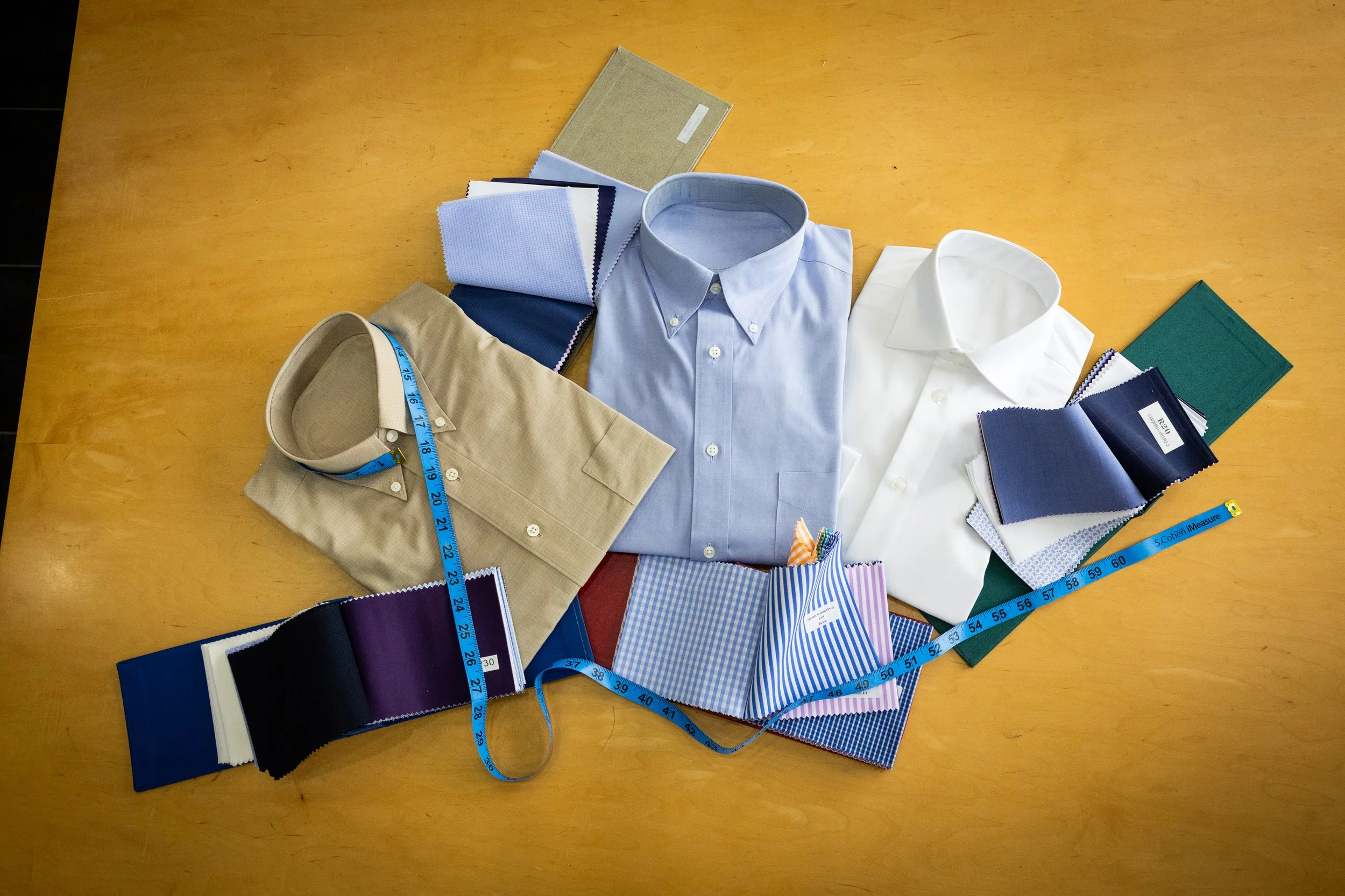 Arranged men's dress shirts in light blue, white, and beige with fabric swatches, measuring tapes, and color samples on a wooden table.