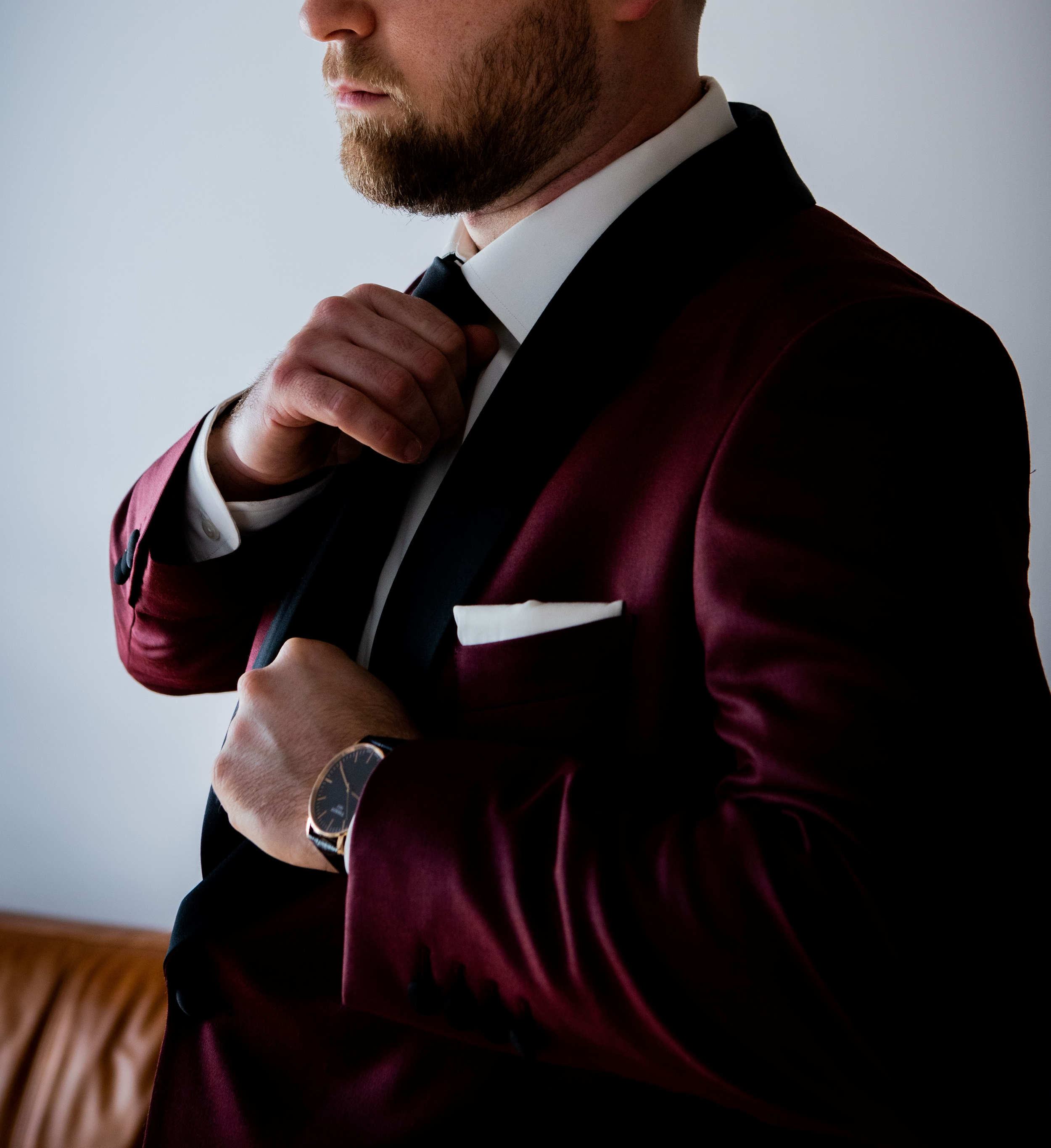 Man adjusting his bow tie, dressed in a burgundy tuxedo with black lapel, white shirt, and cufflinks, standing indoors.