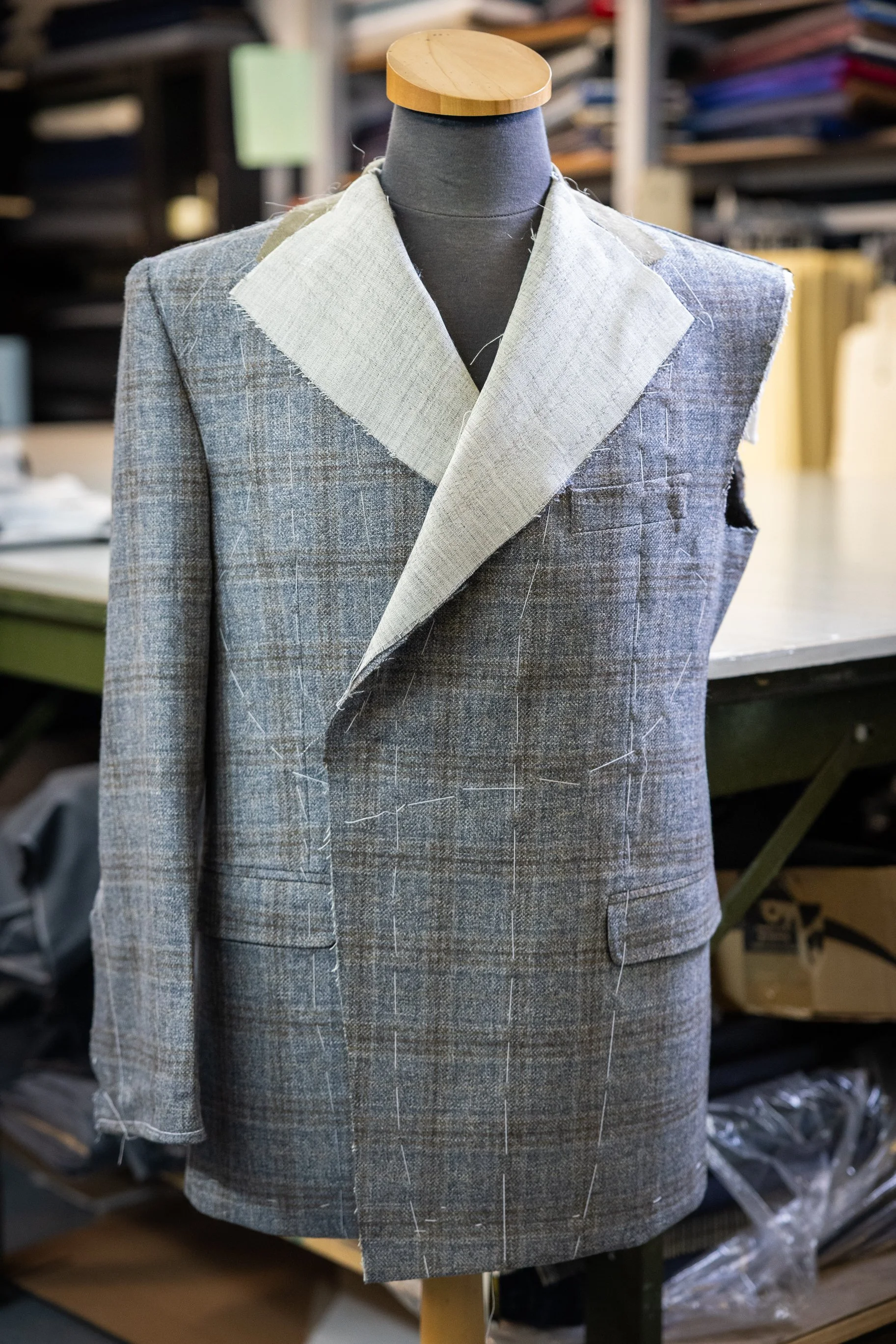 A mannequin wearing a grey plaid suit jacket with an unfinished collar in a tailoring workshop.